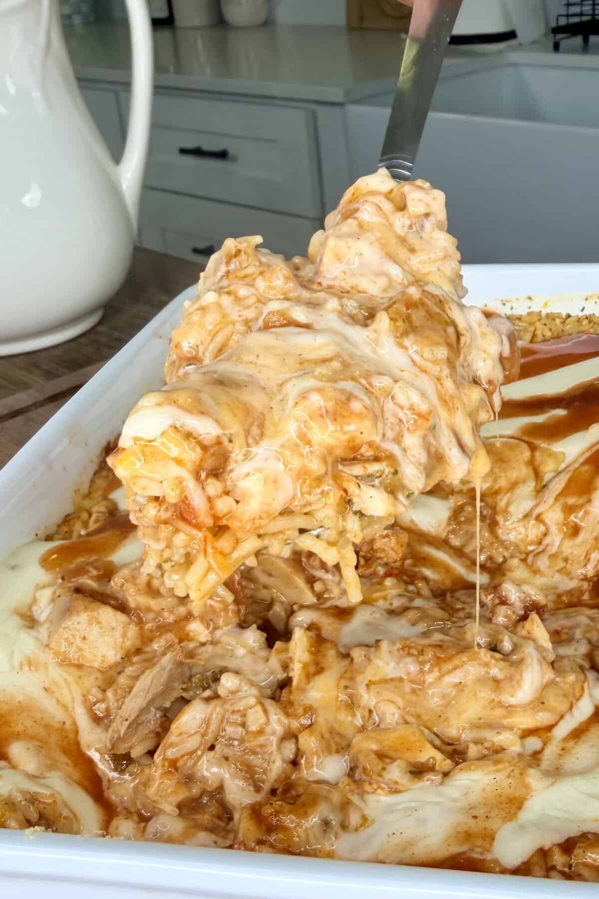 A serving spoon lifts a cheesy, saucy casserole from a white baking dish, revealing layers of melted cheese, rice, and shredded meat, with sauce dripping down.