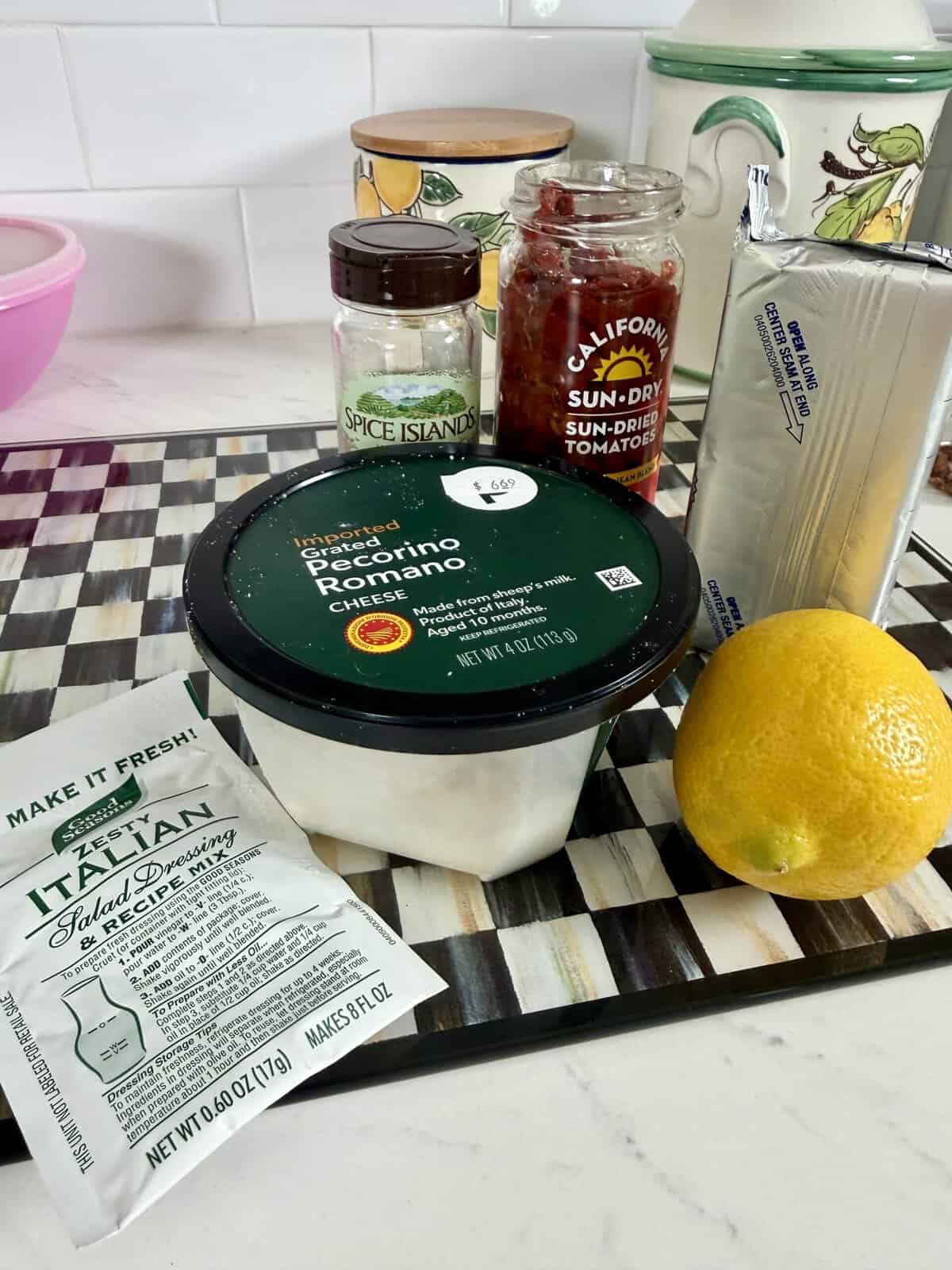 A cutting board holds a lemon, Pecorino Romano cheese, cream cheese, sun-dried tomatoes, Italian dressing mix, and a small spice jar. Kitchen items are visible in the background.