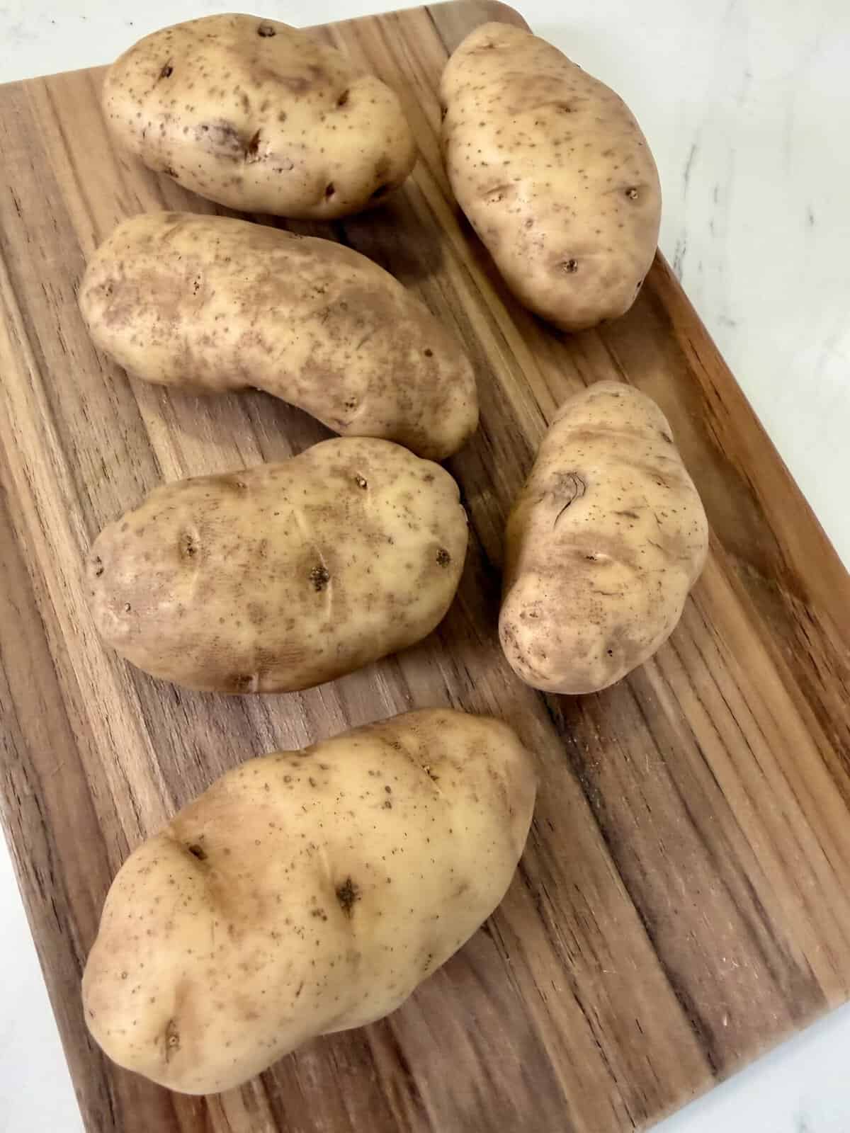Six unpeeled potatoes are placed on a rectangular wooden cutting board, which sits on a white surface. The potatoes have light brown skin with some speckles and small eyes.