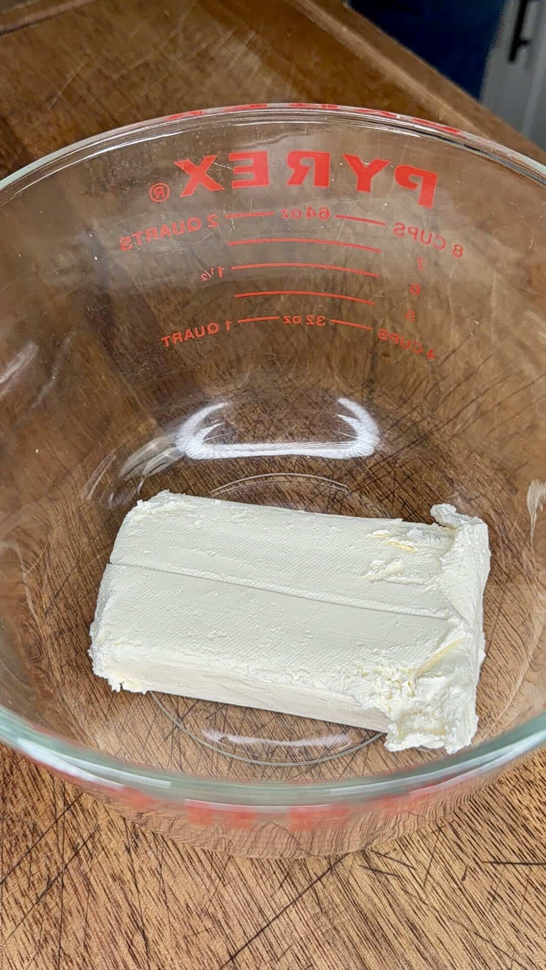 A block of cream cheese in a glass Pyrex measuring bowl sits on a wooden countertop.