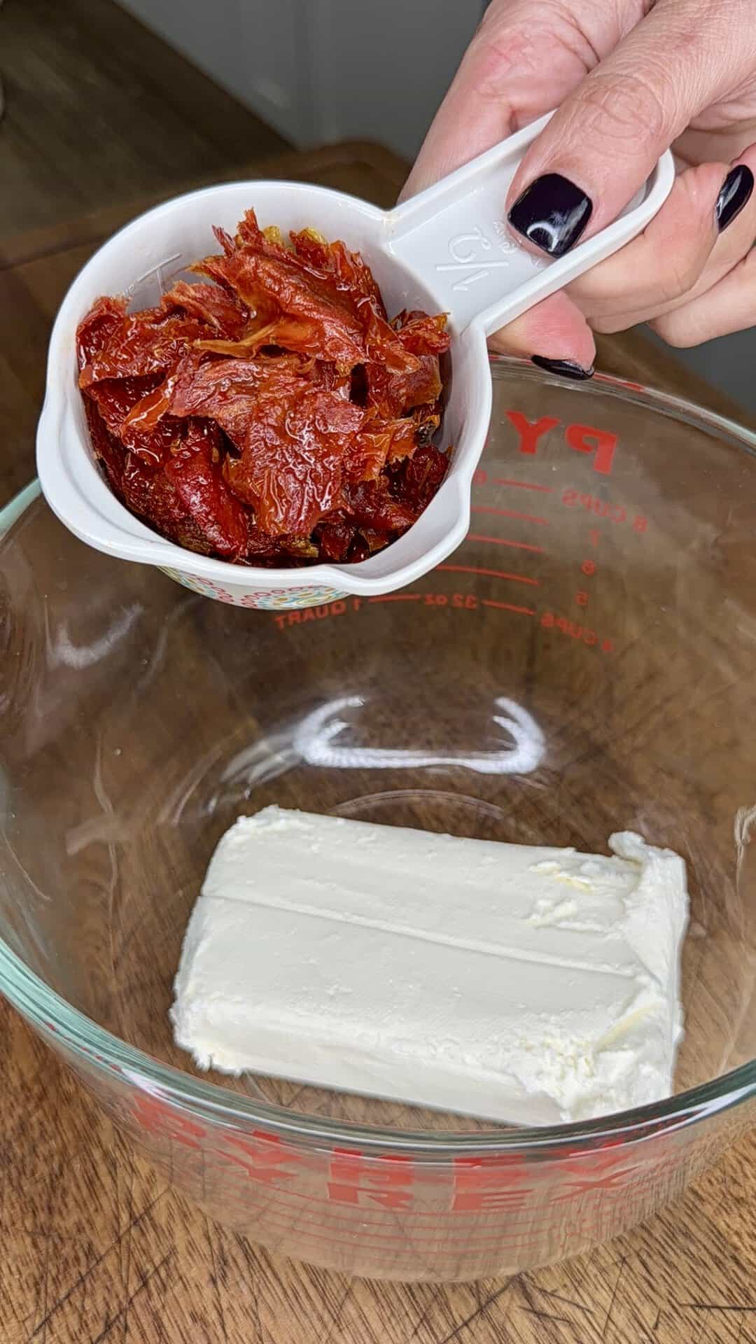 A hand holds a white measuring cup with chopped sun-dried tomatoes above a glass bowl containing a block of cream cheese on a wooden surface.