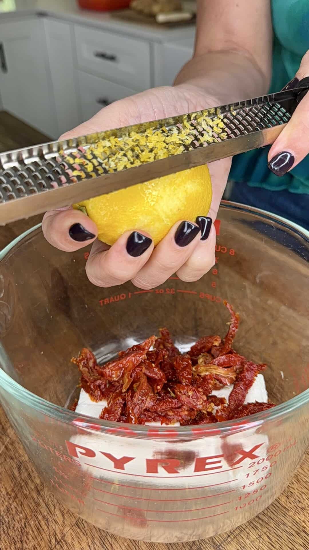 A person grates lemon zest over a glass measuring cup filled with sun-dried tomatoes and a white block, likely cheese or tofu.