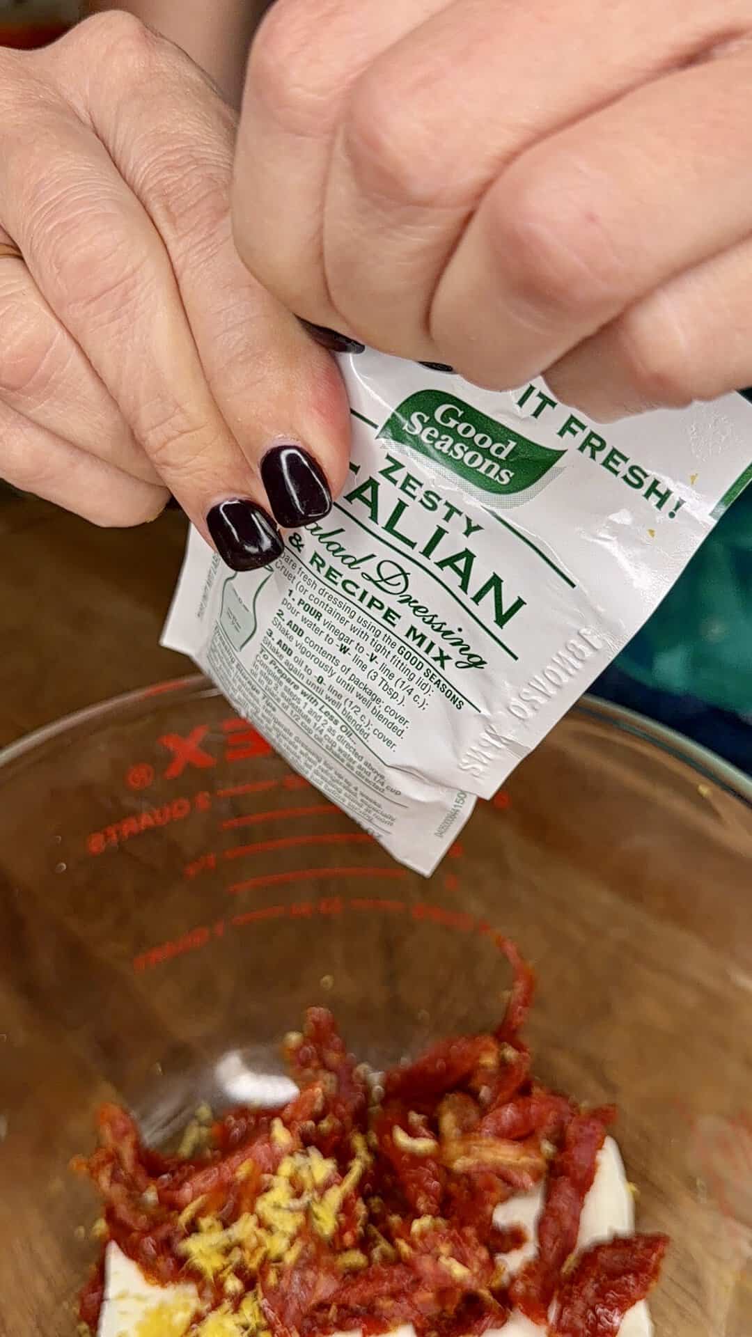 A person with dark nail polish pours Good Seasons Zesty Italian dressing mix into a glass bowl containing sun-dried tomatoes, garlic, and lemon zest.