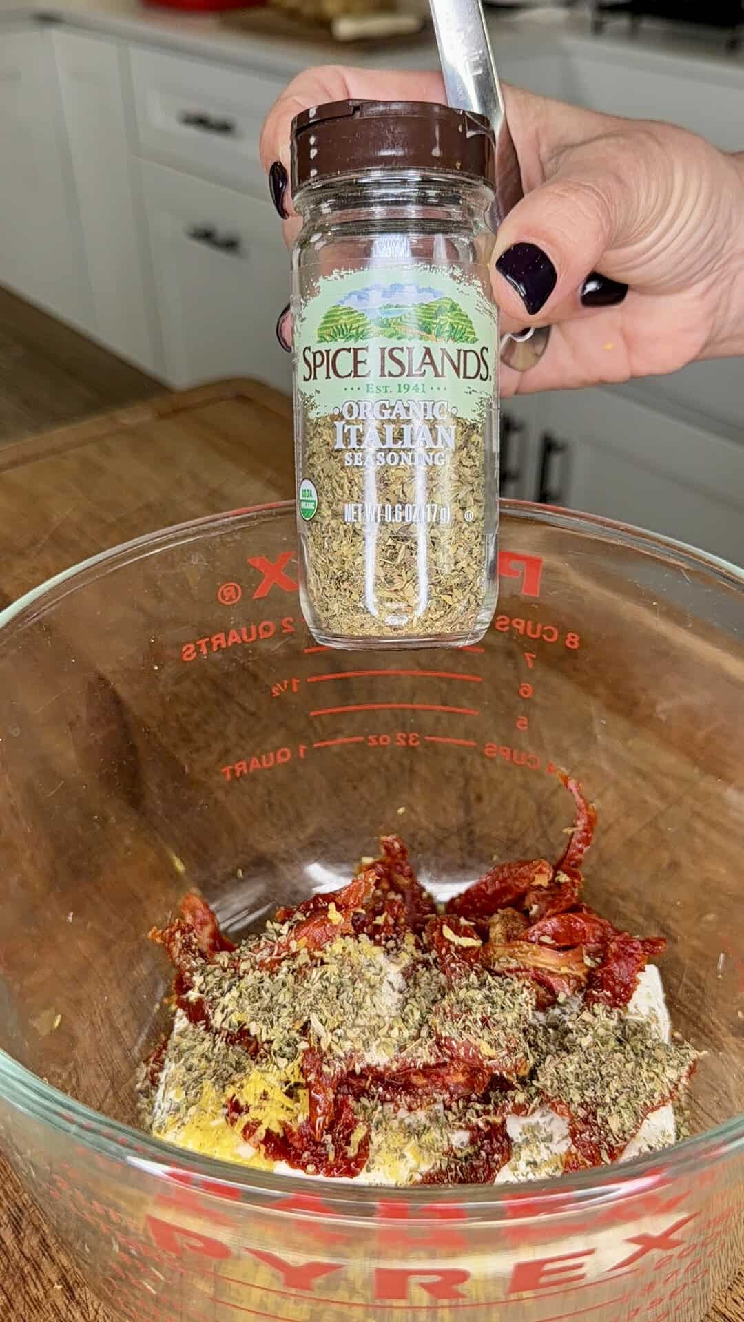 A hand holds a jar of Spice Islands Italian seasoning over a glass bowl filled with herbs, spices, and tomato paste in a kitchen setting.