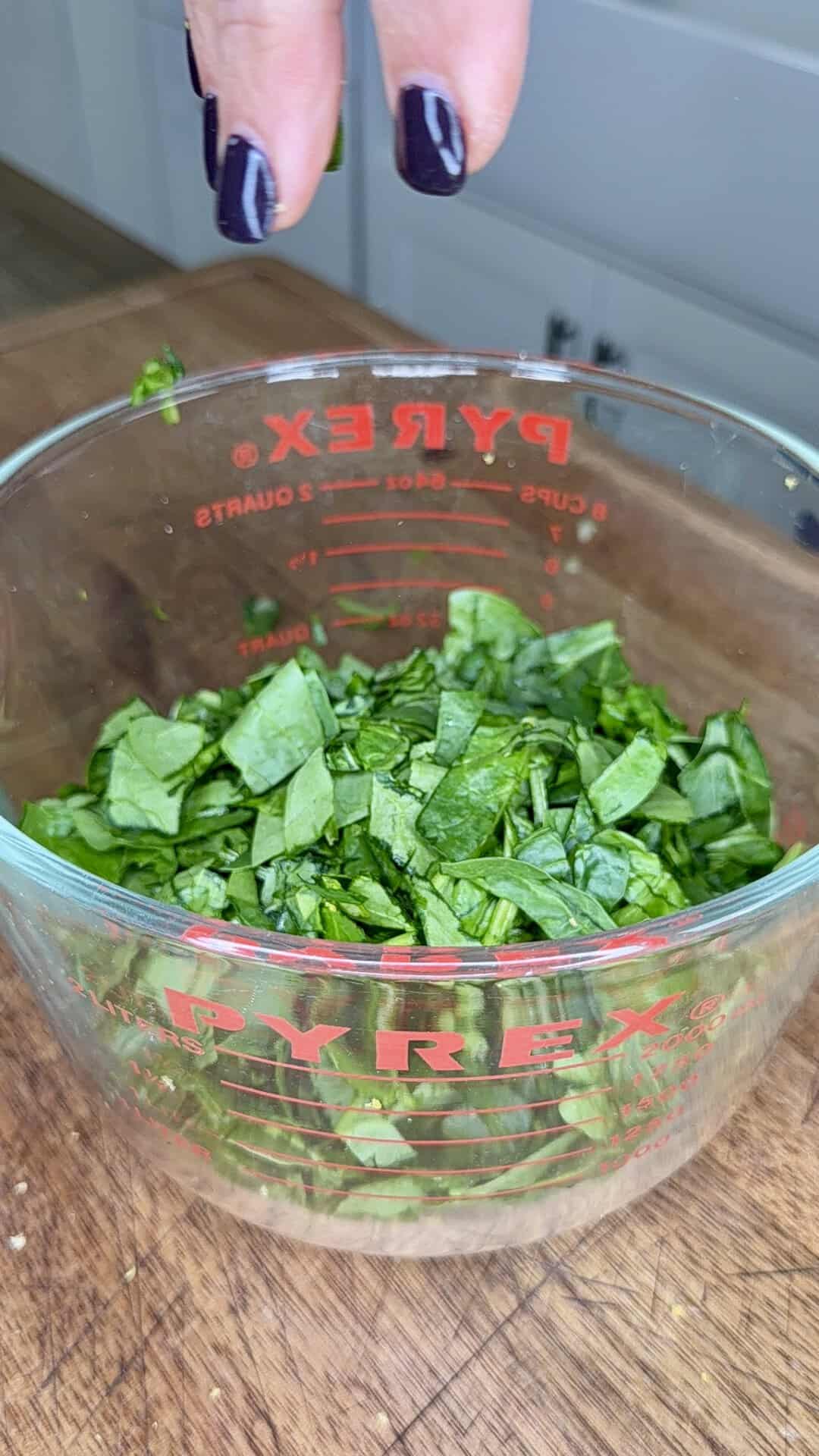 A hand with dark nail polish sprinkles chopped greens into a Pyrex measuring cup on a wooden surface in a kitchen.