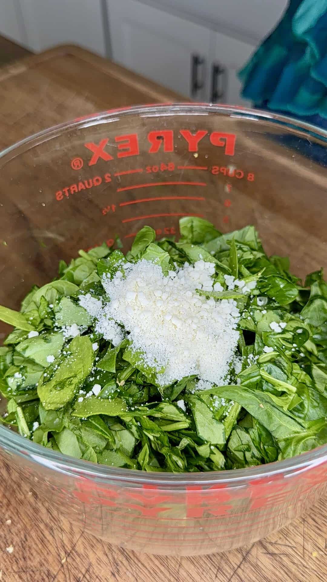 Chopped spinach and grated parmesan cheese in a glass Pyrex measuring cup on a wooden countertop, kitchen cabinets in the background.