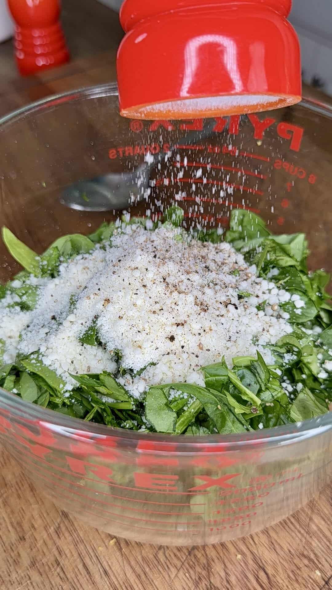 Ground pepper falls from a red grinder onto chopped spinach and grated cheese in a glass Pyrex measuring cup on a wooden surface.
