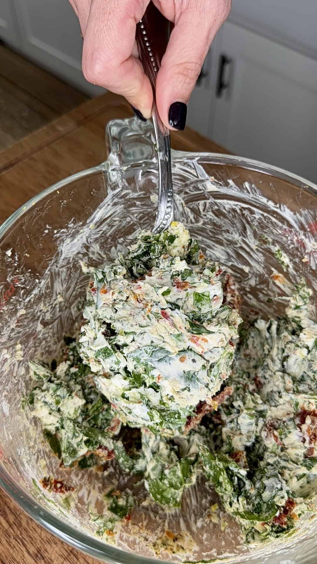 A hand with dark nail polish holds a spoon mixing creamy spinach dip with visible greens and red bits in a glass bowl on a wooden countertop.