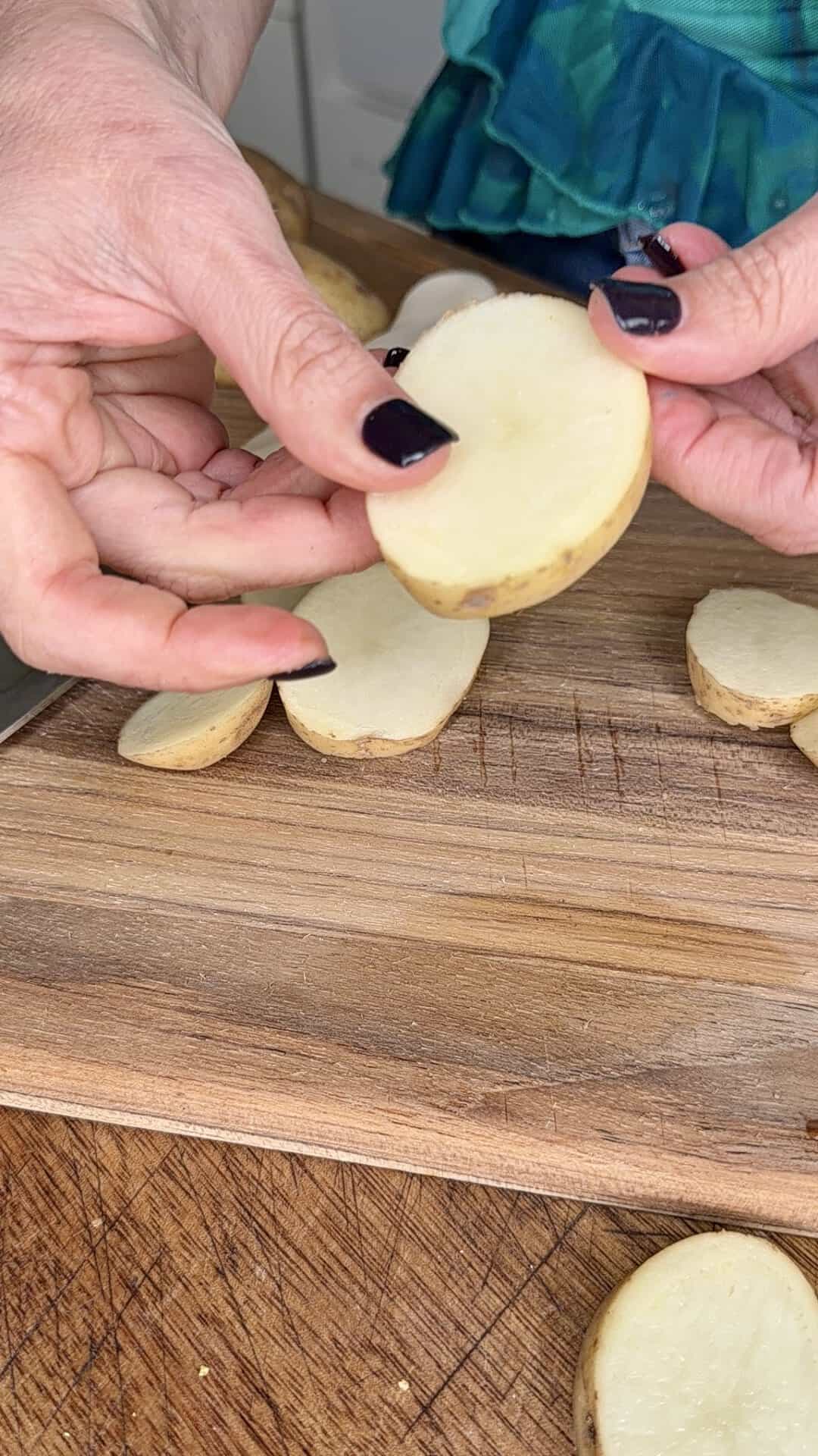 Close-up of hands holding a raw potato slice over a wooden cutting board, with additional potato slices and a person wearing a green top visible in the background.