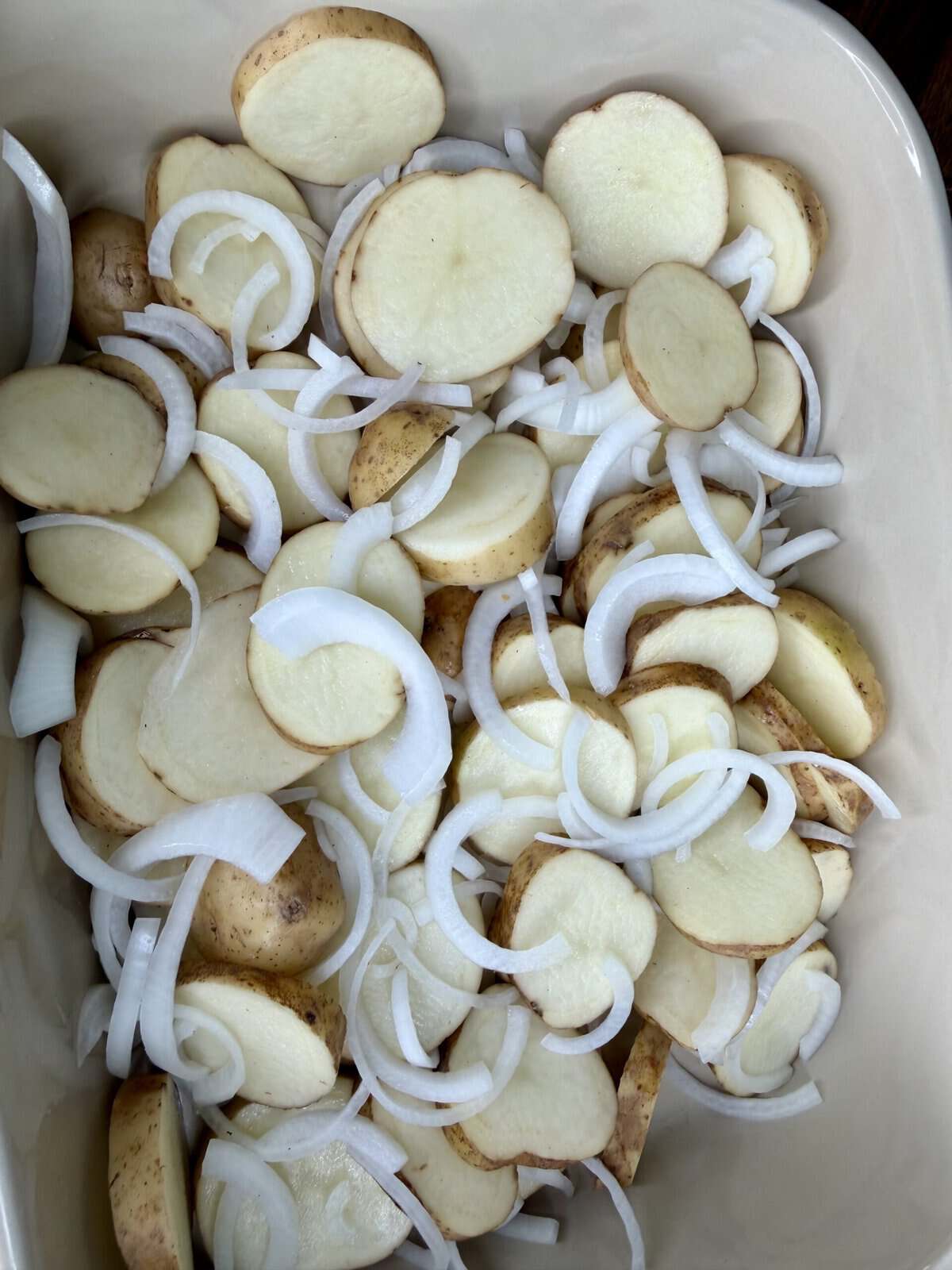 Sliced potatoes and raw onion strips are arranged in a beige baking dish, ready for cooking.