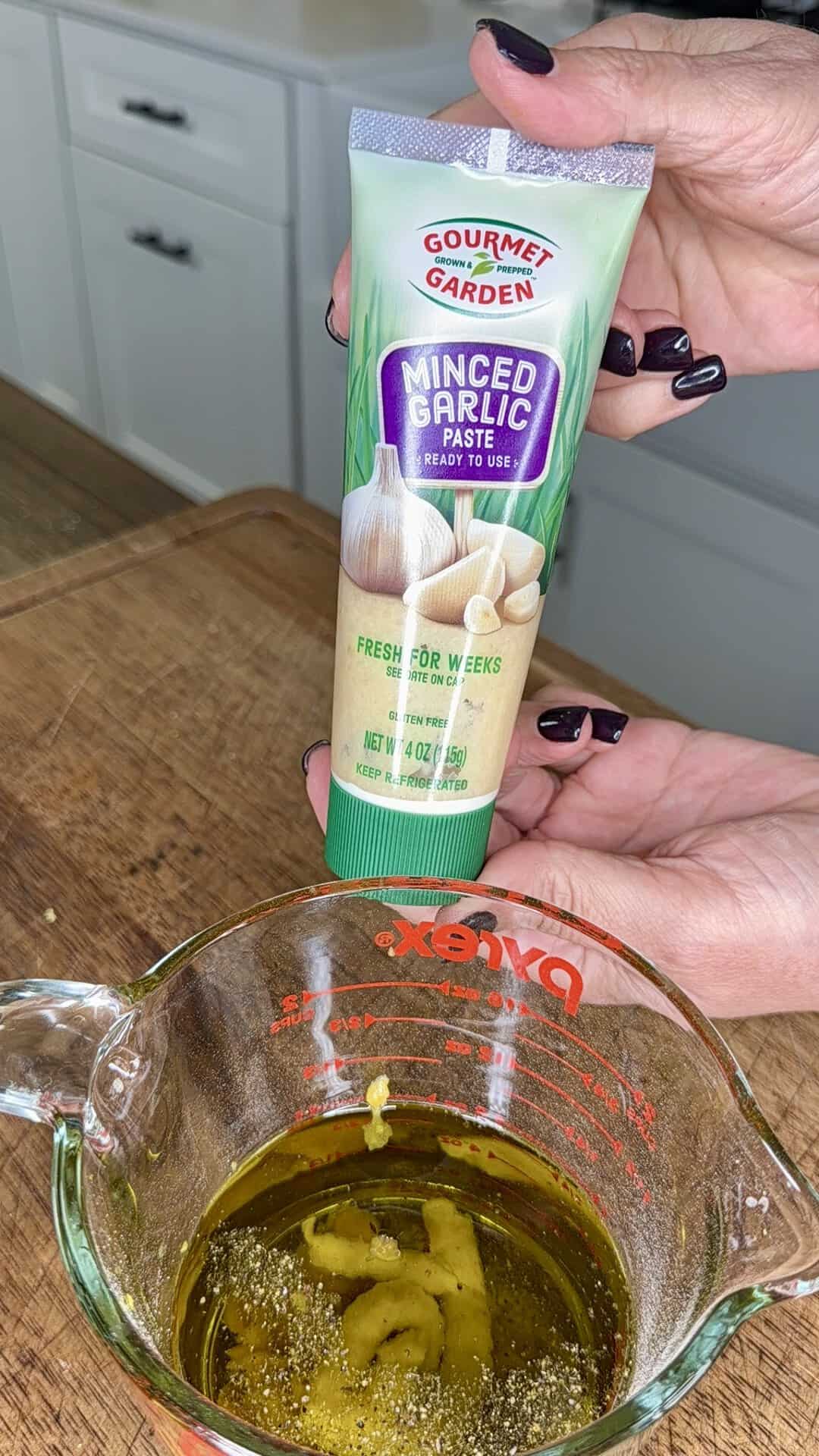 Hands hold a tube of Gourmet Garden minced garlic paste above a glass measuring cup filled with olive oil and seasonings on a kitchen counter.