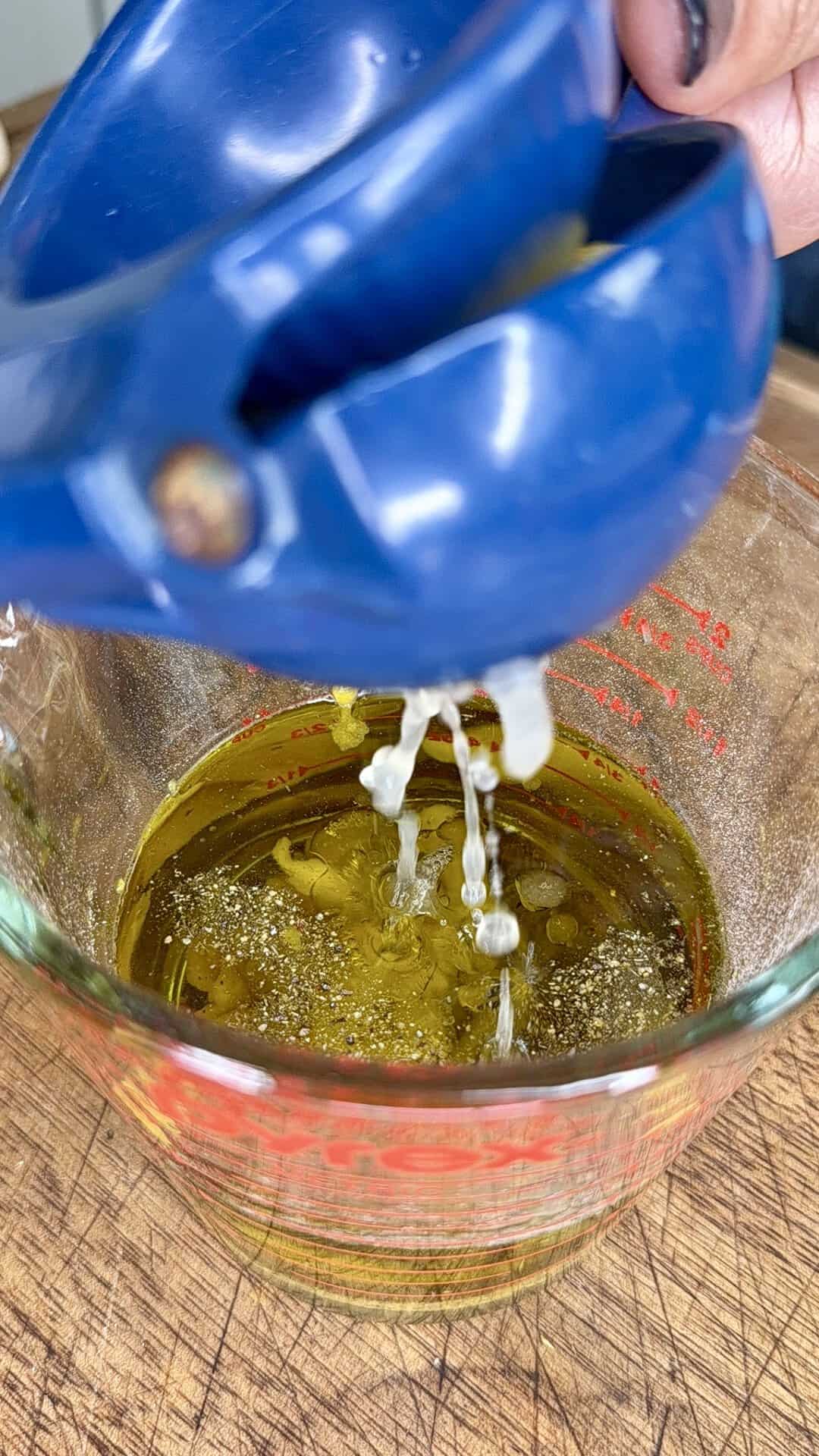 Lemon juice being squeezed from a blue citrus press into a glass measuring cup filled with olive oil, on a wooden surface.