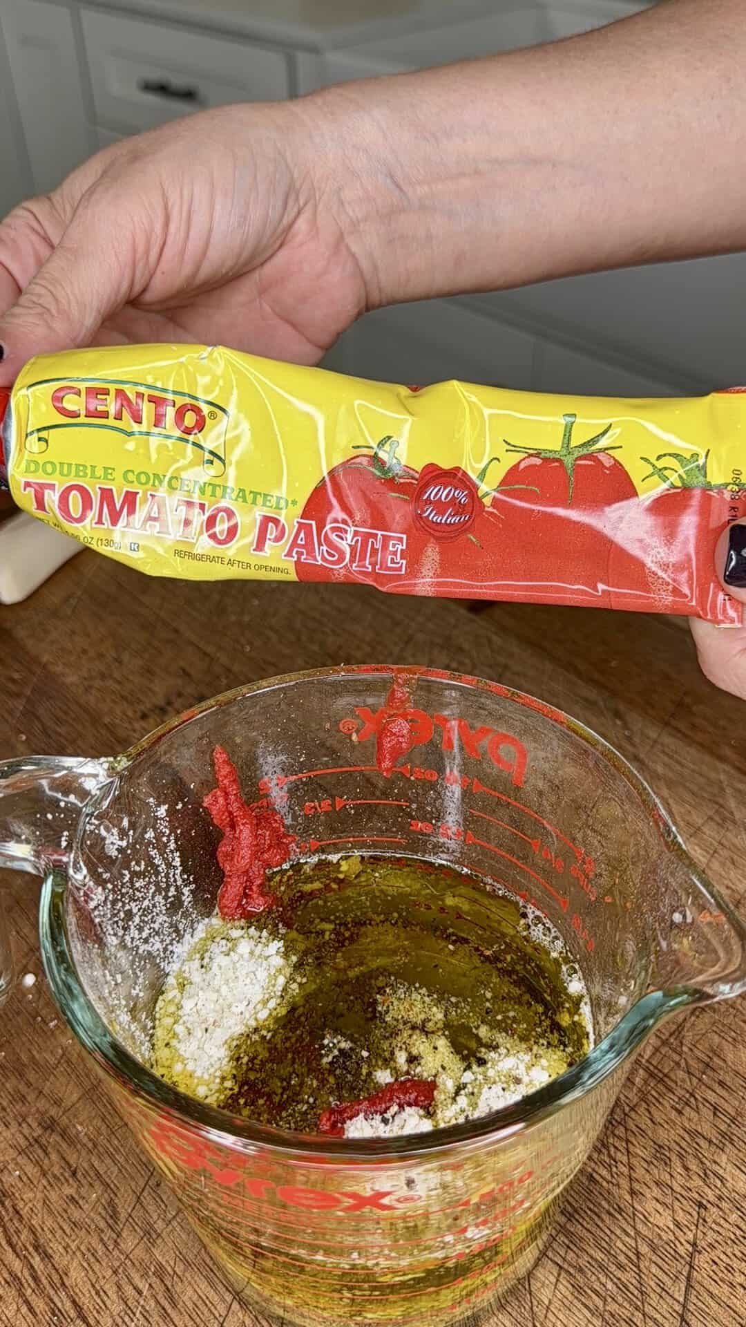A hand holds a tube of Cento tomato paste above a glass measuring cup filled with oil, spices, and some tomato paste on a wooden surface.