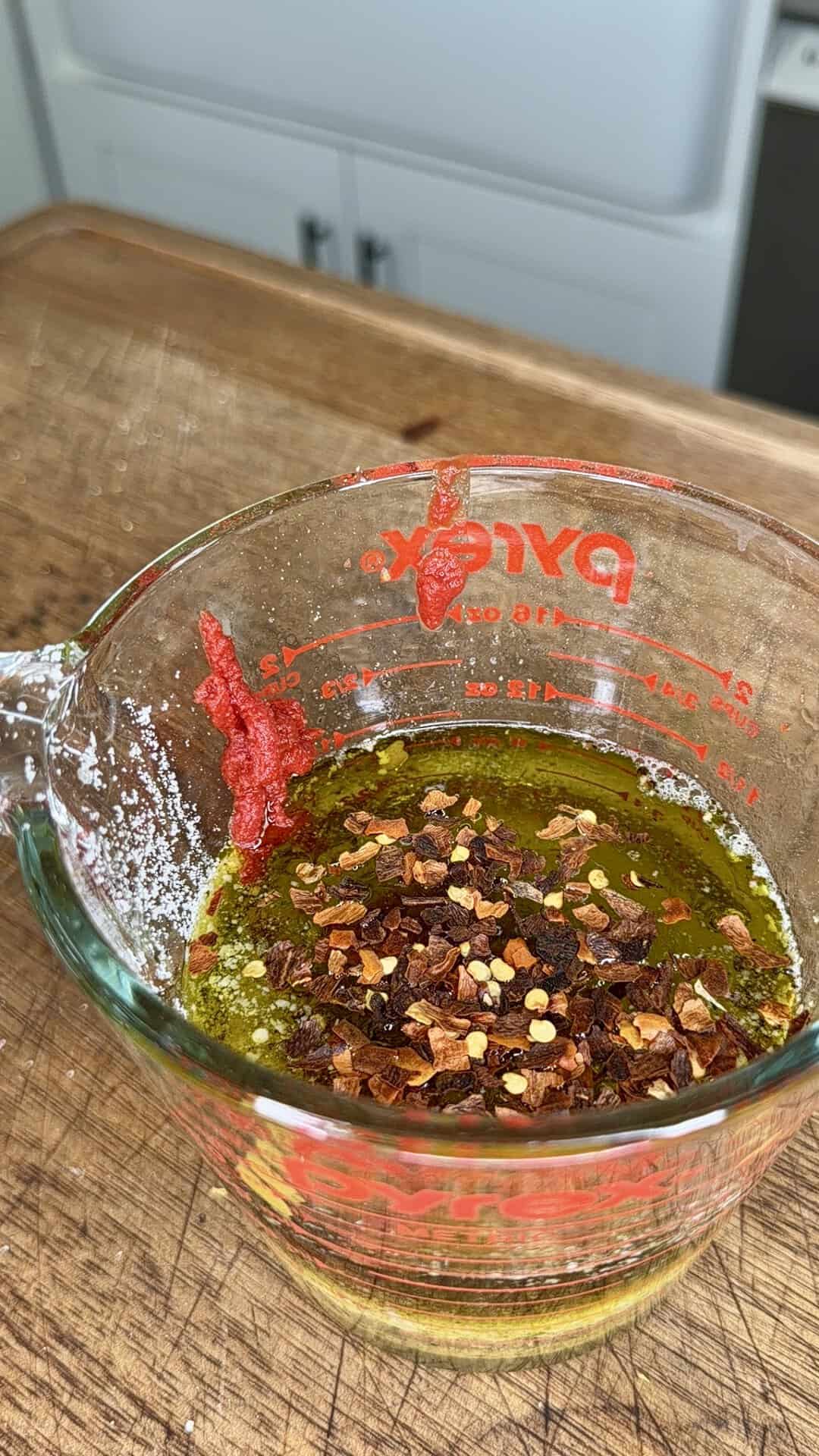 A glass measuring cup with olive oil, chili flakes, and red paste sits on a wooden counter in a kitchen.