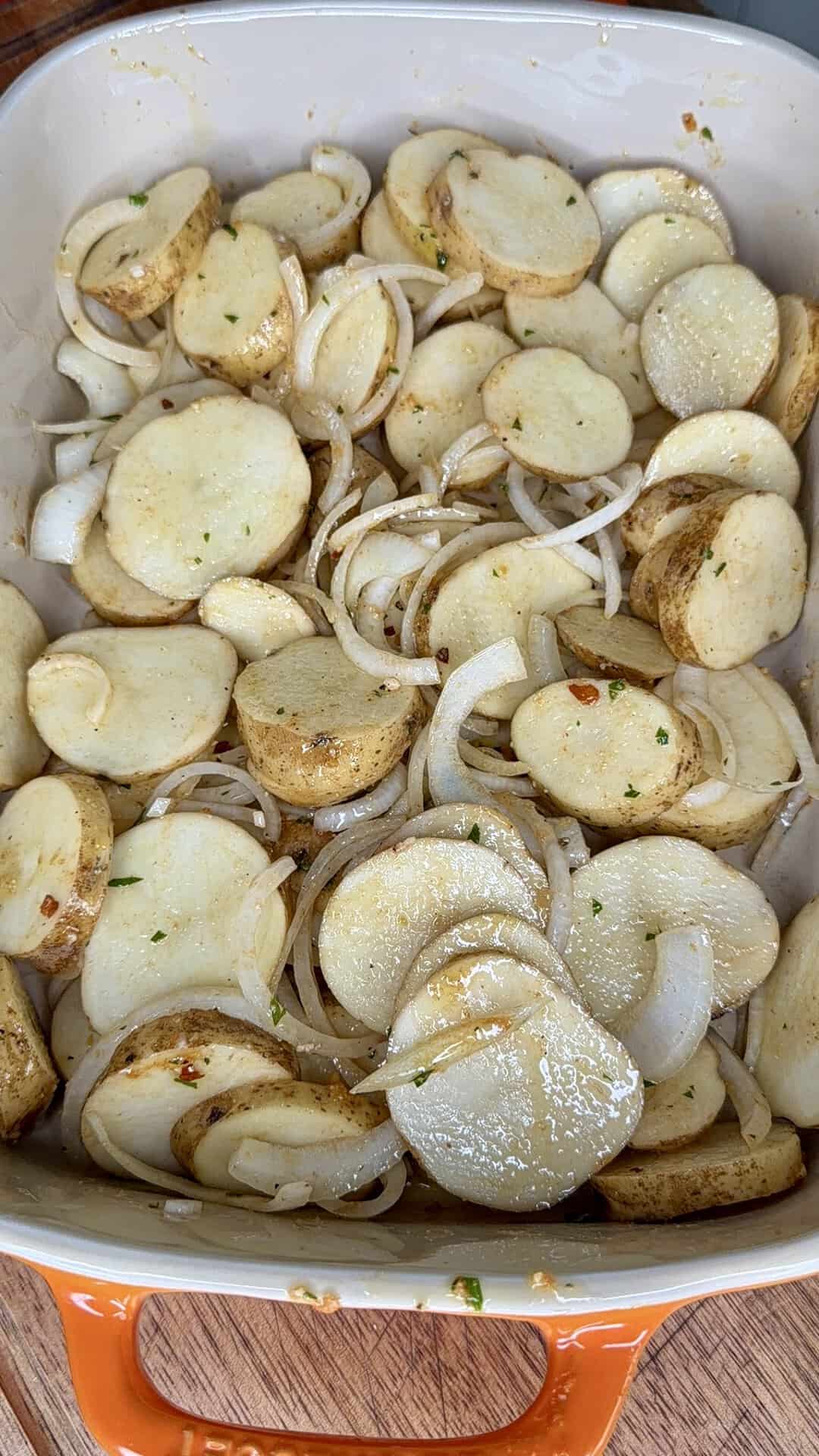 Sliced potatoes and onions coated in seasoning are arranged in a rectangular orange baking dish, ready to be cooked.