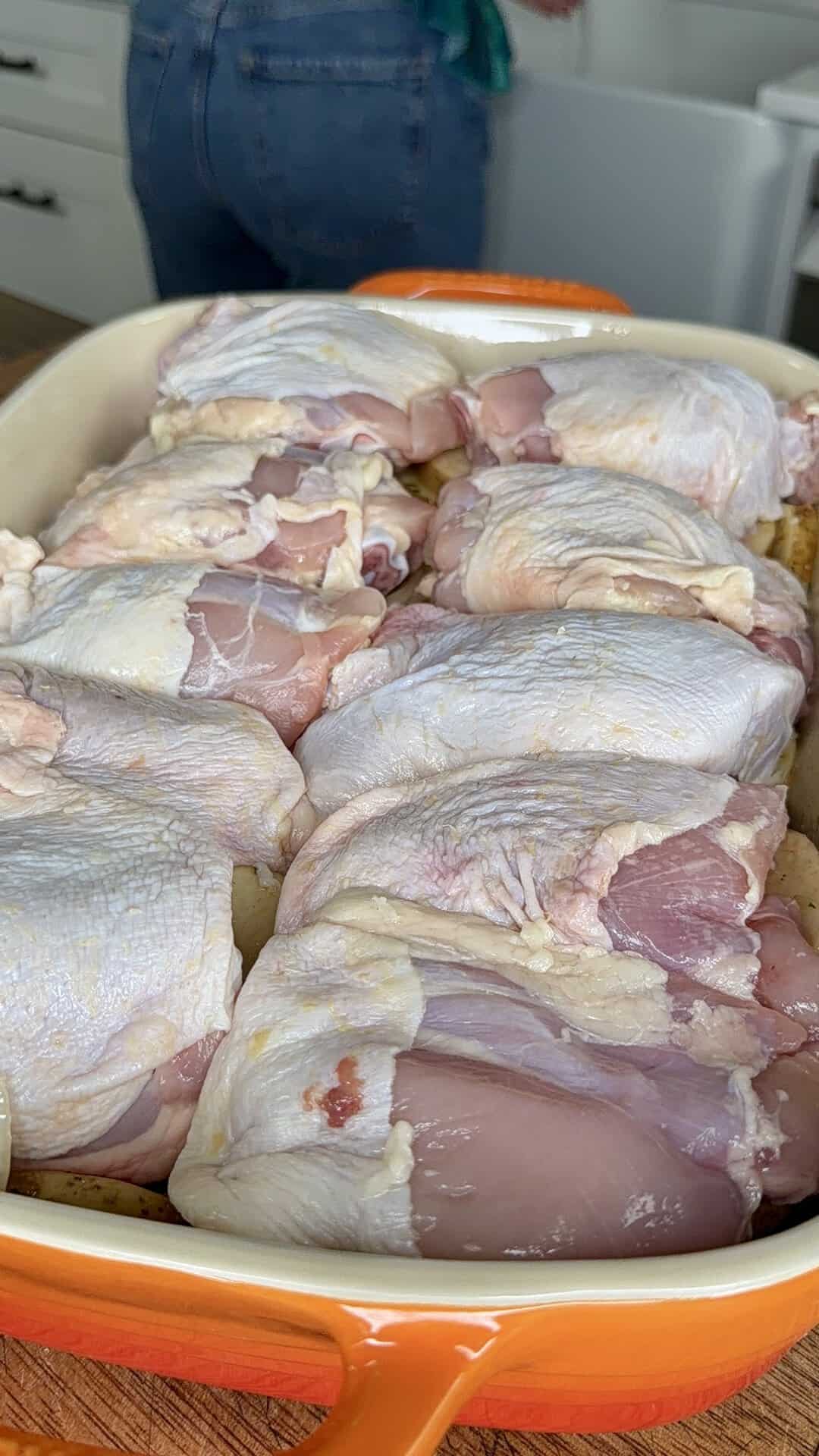 Raw chicken thighs arranged in a casserole dish, ready for cooking. A person is visible in the background near a kitchen counter and cabinets.