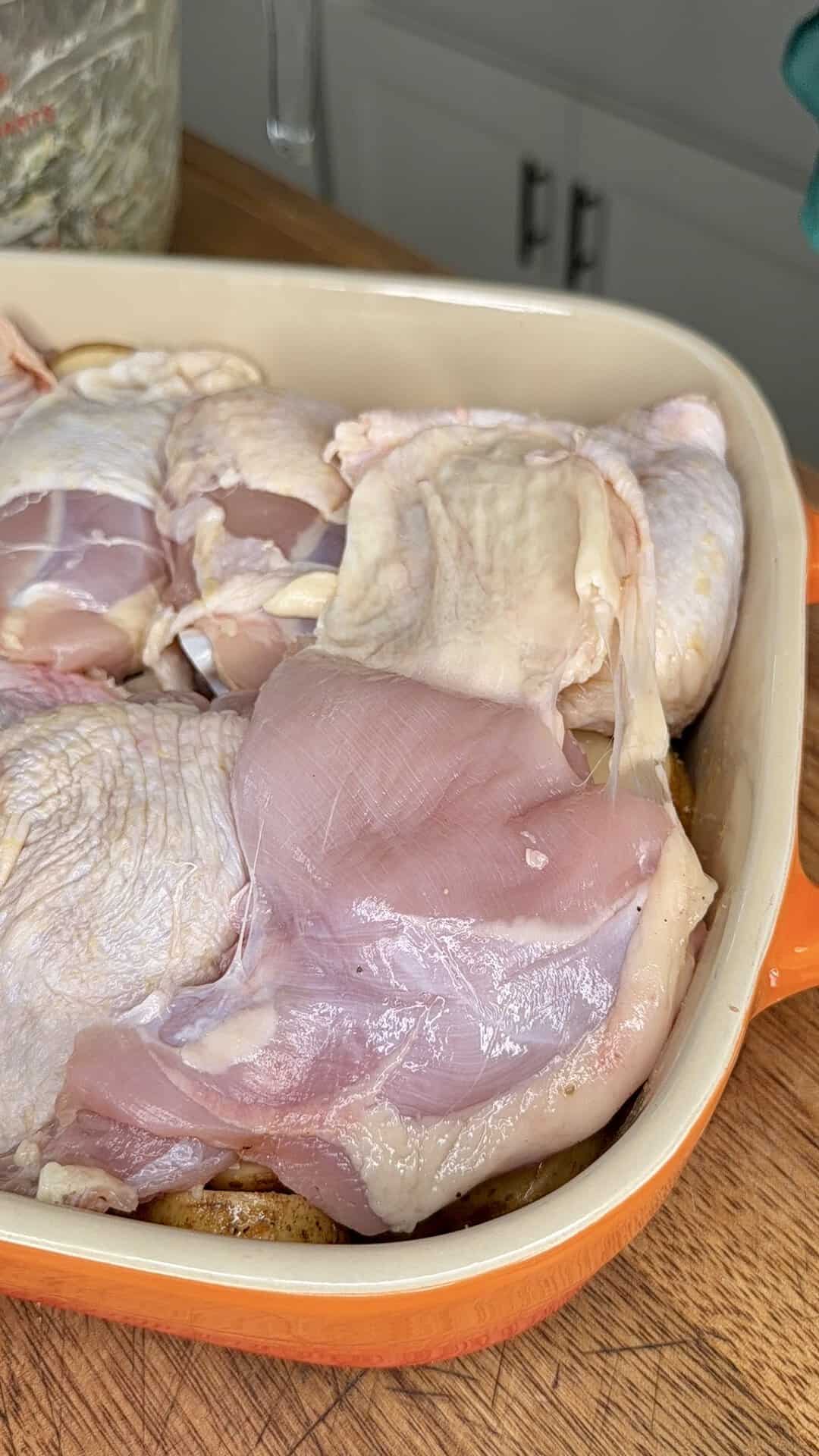 Raw chicken pieces arranged in an orange baking dish, placed on a wooden countertop in a kitchen setting.