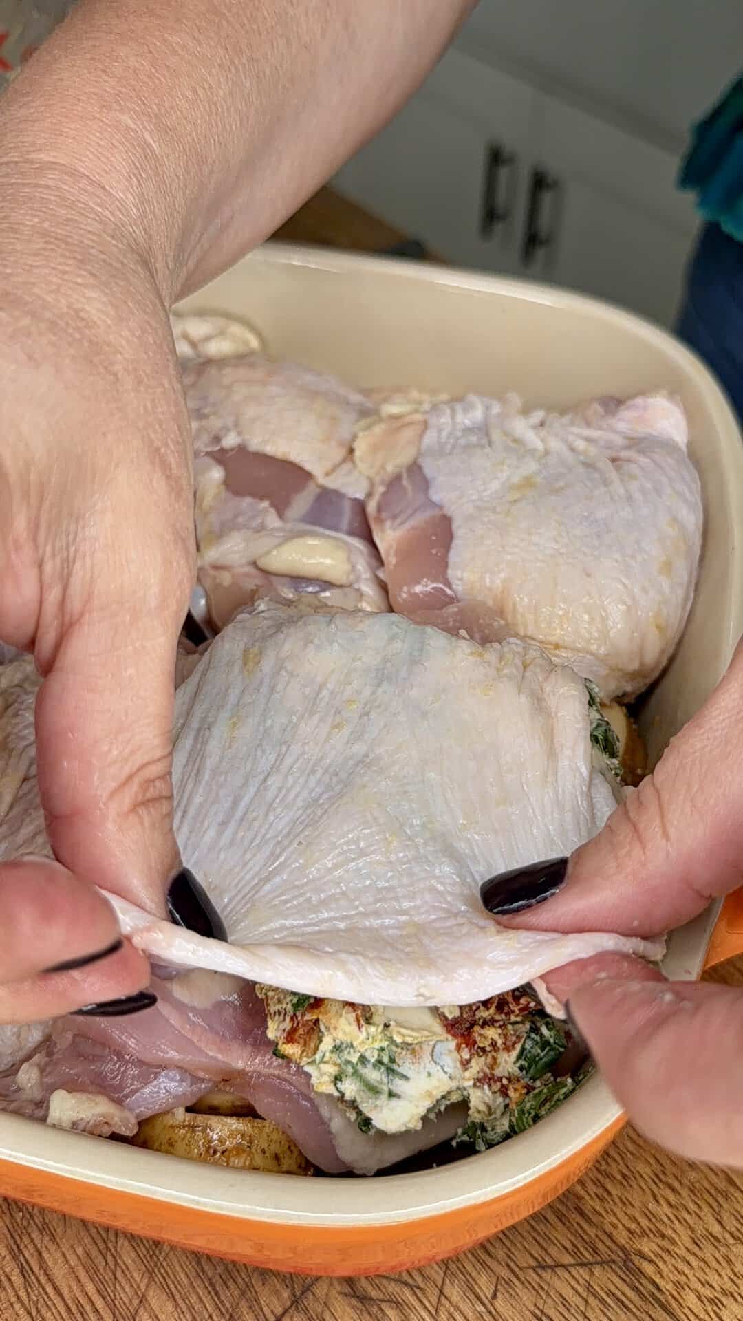 Hands stuffing chicken thighs with a spinach and cheese mixture in a baking dish, preparing them for cooking on a wooden surface in a kitchen.