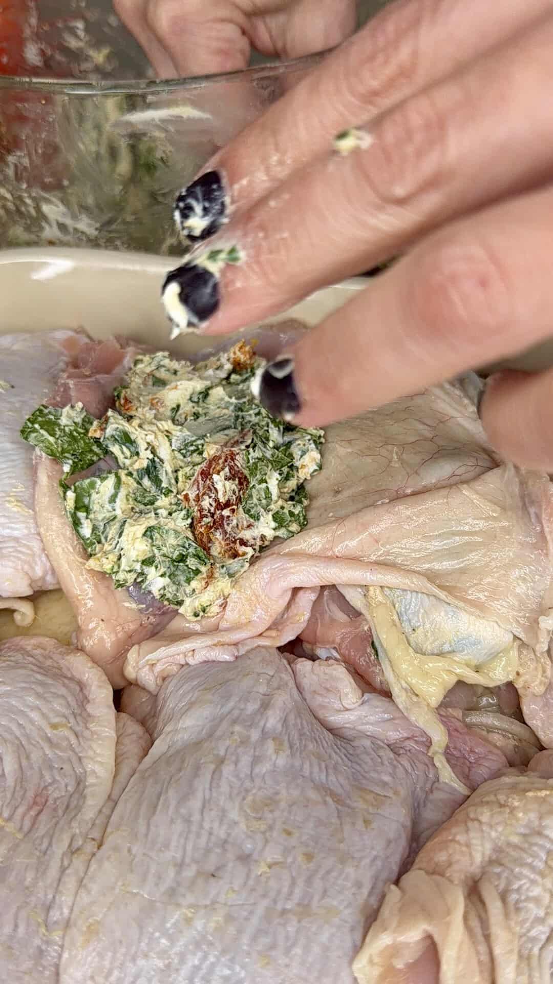 A hand with painted nails spreads a herb and butter mixture onto raw chicken pieces in a dish.