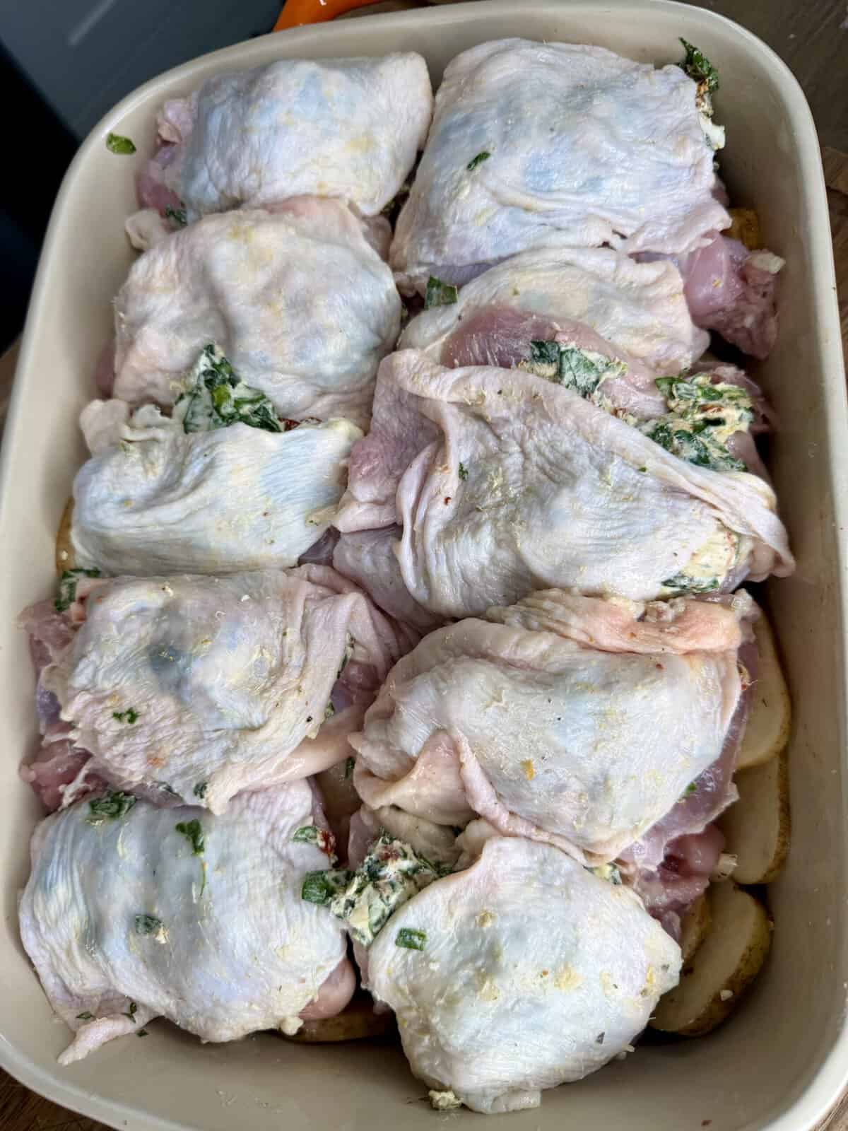 Uncooked chicken thighs with skin, some stuffed with a green and white herb mixture, arranged in a baking dish over sliced potatoes.