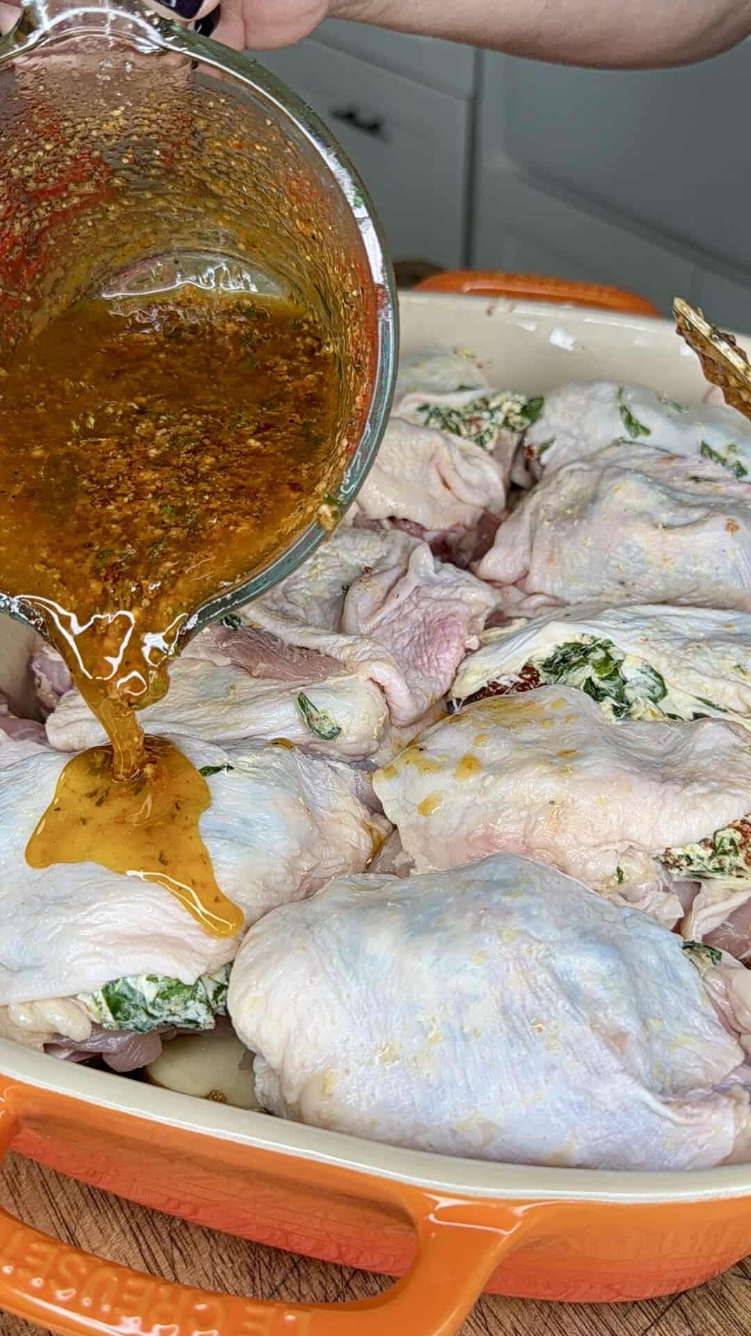 A hand pours a seasoned marinade over raw chicken pieces stuffed with a green herb mixture, arranged in an orange baking dish on a kitchen counter.