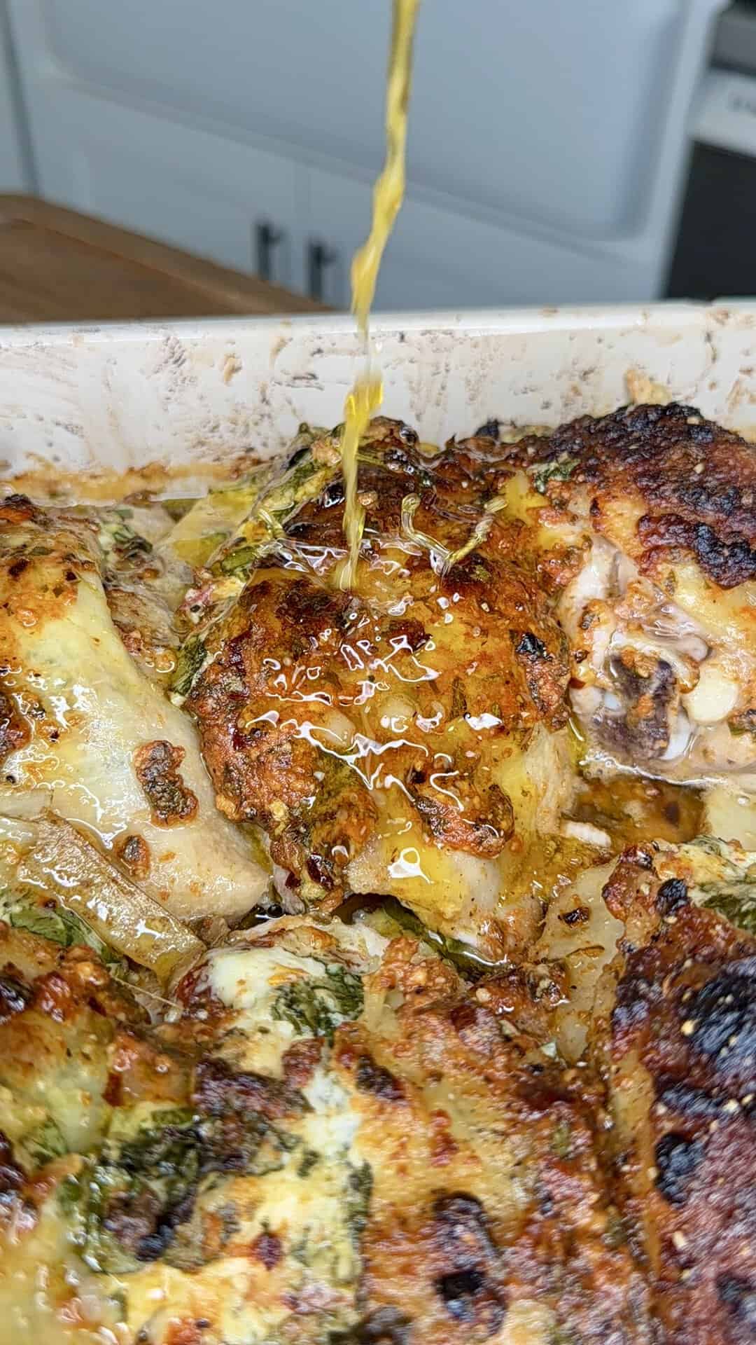 Golden liquid being poured over baked, seasoned chicken pieces in a dish, highlighting crispy skin and herbs, with a kitchen background slightly visible.