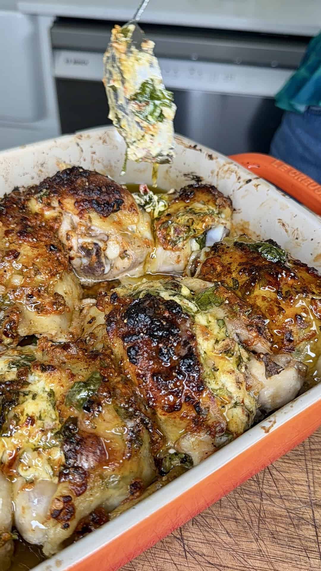 Golden-brown roasted chicken pieces in an orange baking dish, topped with herbs and melted butter, with a utensil scooping herbed butter onto the chicken.