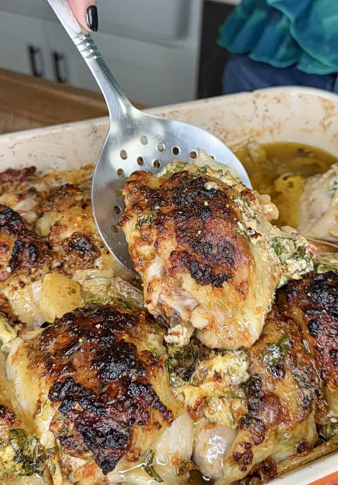 A hand holds a slotted spoon, lifting a golden-brown baked chicken thigh with herbs and cheese from a baking dish filled with more chicken.