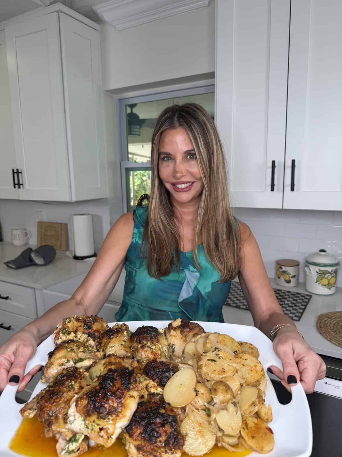 A woman in a green top smiles while holding a large plate of roasted chicken and potatoes in a bright, modern kitchen.