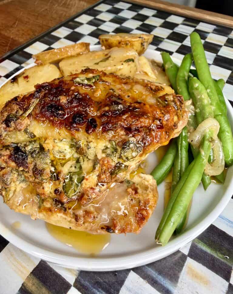 A plate with baked chicken topped with herbs, roasted potato slices, and sautéed green beans with onions, on a checkered placemat.