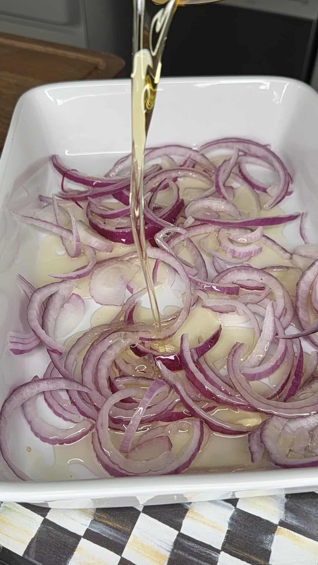 Sliced red onions in a white dish as a stream of oil is poured over them, with a checkered cloth partially visible at the bottom of the image.