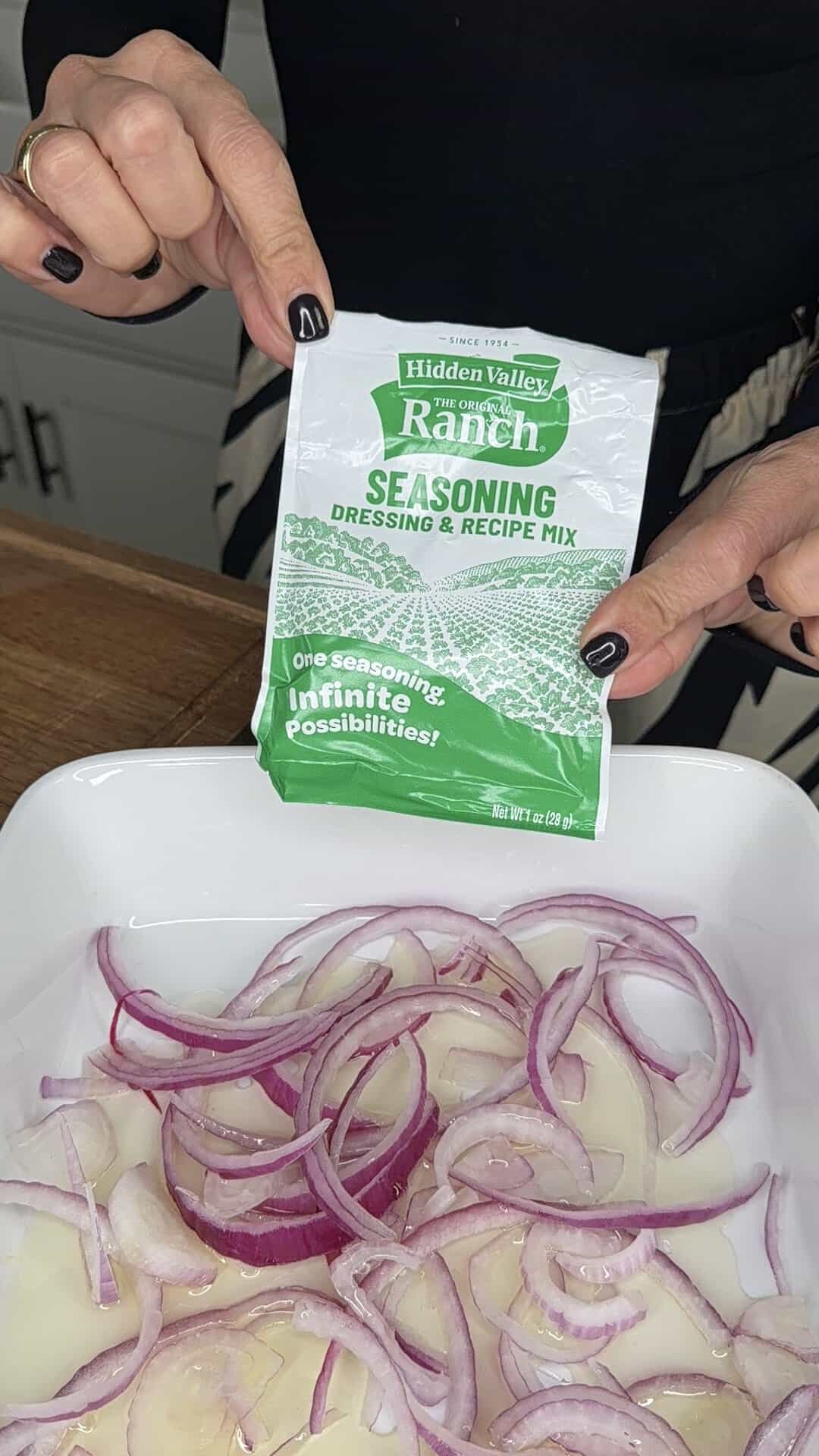 A person holds a packet of Hidden Valley Ranch seasoning above a dish filled with sliced red onions and sliced potatoes.