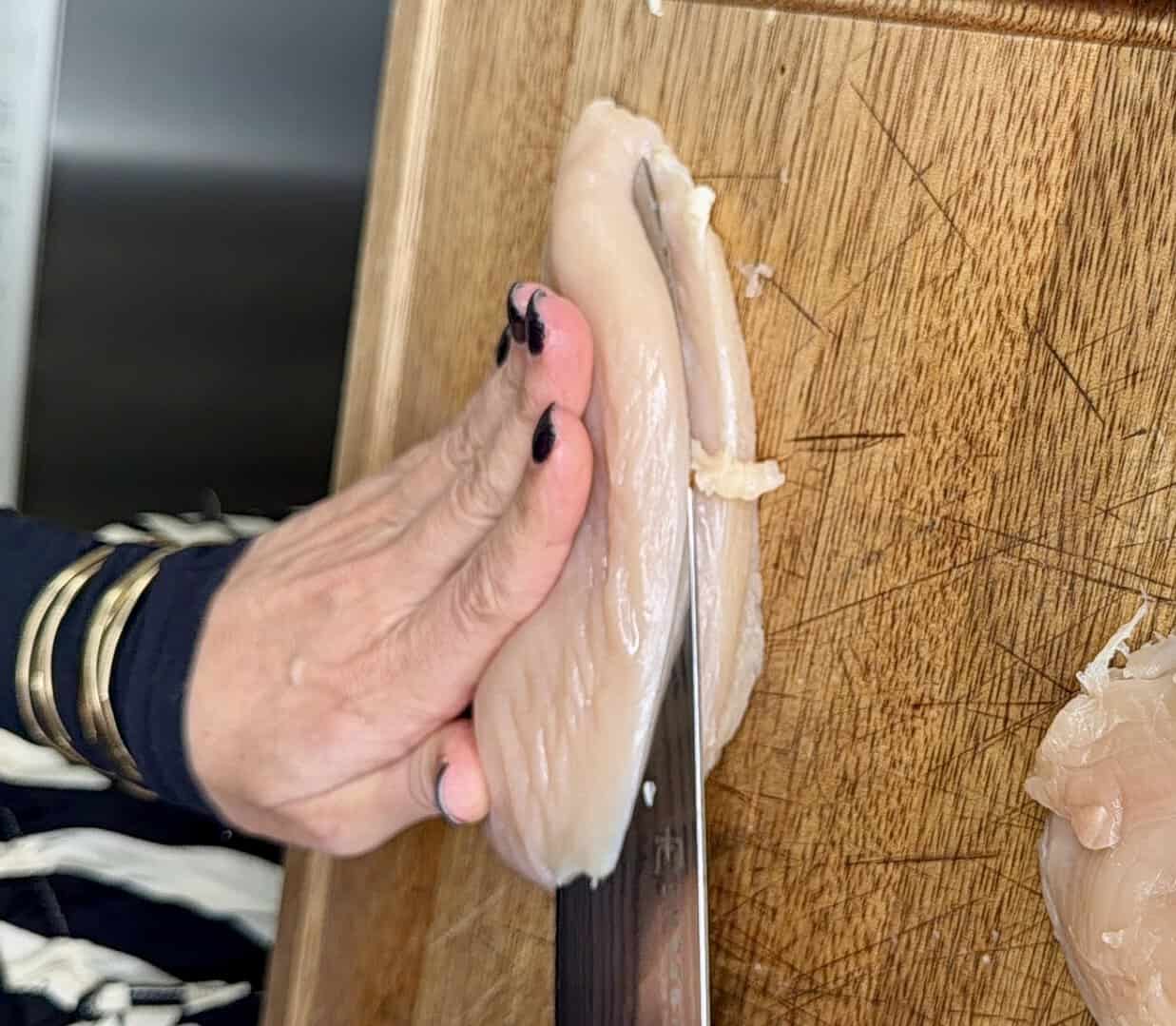 A person with black nail polish slices a raw chicken breast on a wooden cutting board with a large knife.