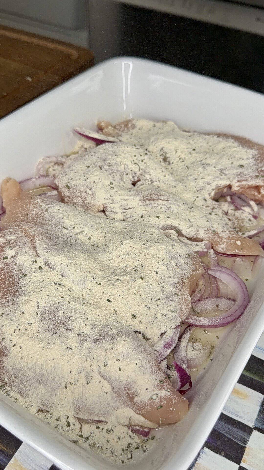 Raw chicken breasts topped with sliced red onions and a layer of powdered seasoning in a white baking dish, ready to be cooked.