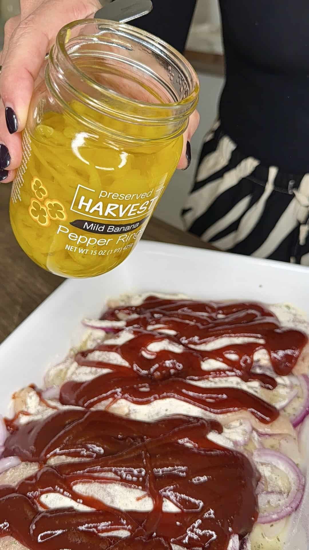 A person holds a jar of mild banana pepper rings above a plate of raw chicken topped with red barbecue sauce and sliced onions, preparing to add the peppers.