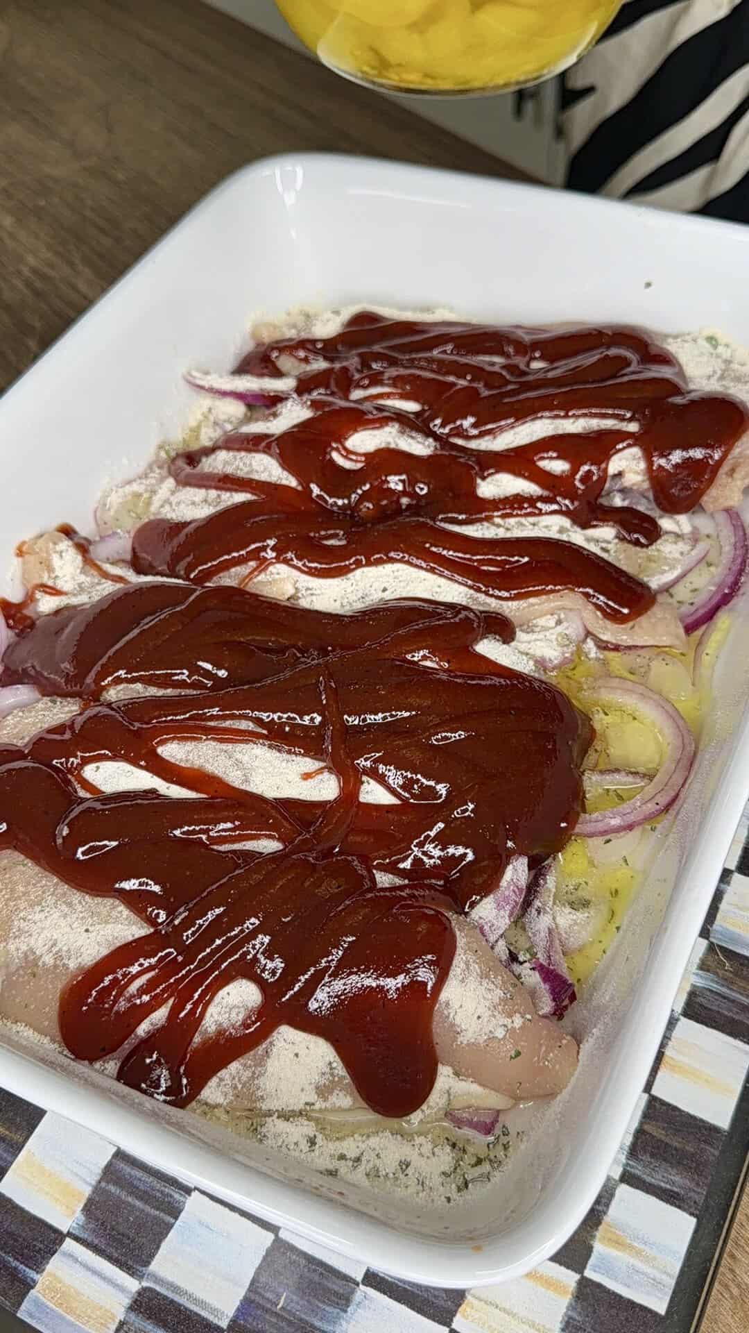 Raw chicken breasts in a white baking dish topped with sliced red onions, seasoning, and a generous layer of barbecue sauce, ready to be cooked.