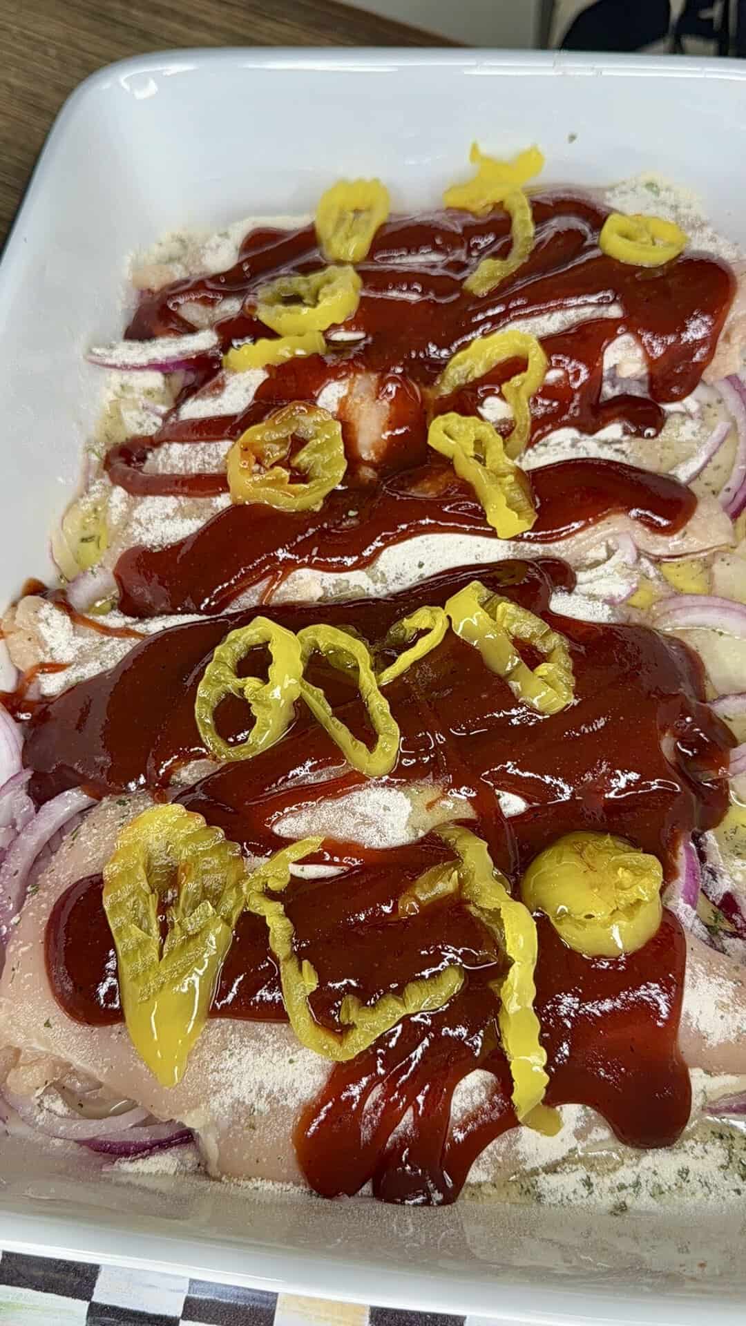 Raw chicken breasts in a white dish topped with sliced red onions, drizzled with barbecue sauce, and garnished with yellow banana pepper rings, ready to be baked.