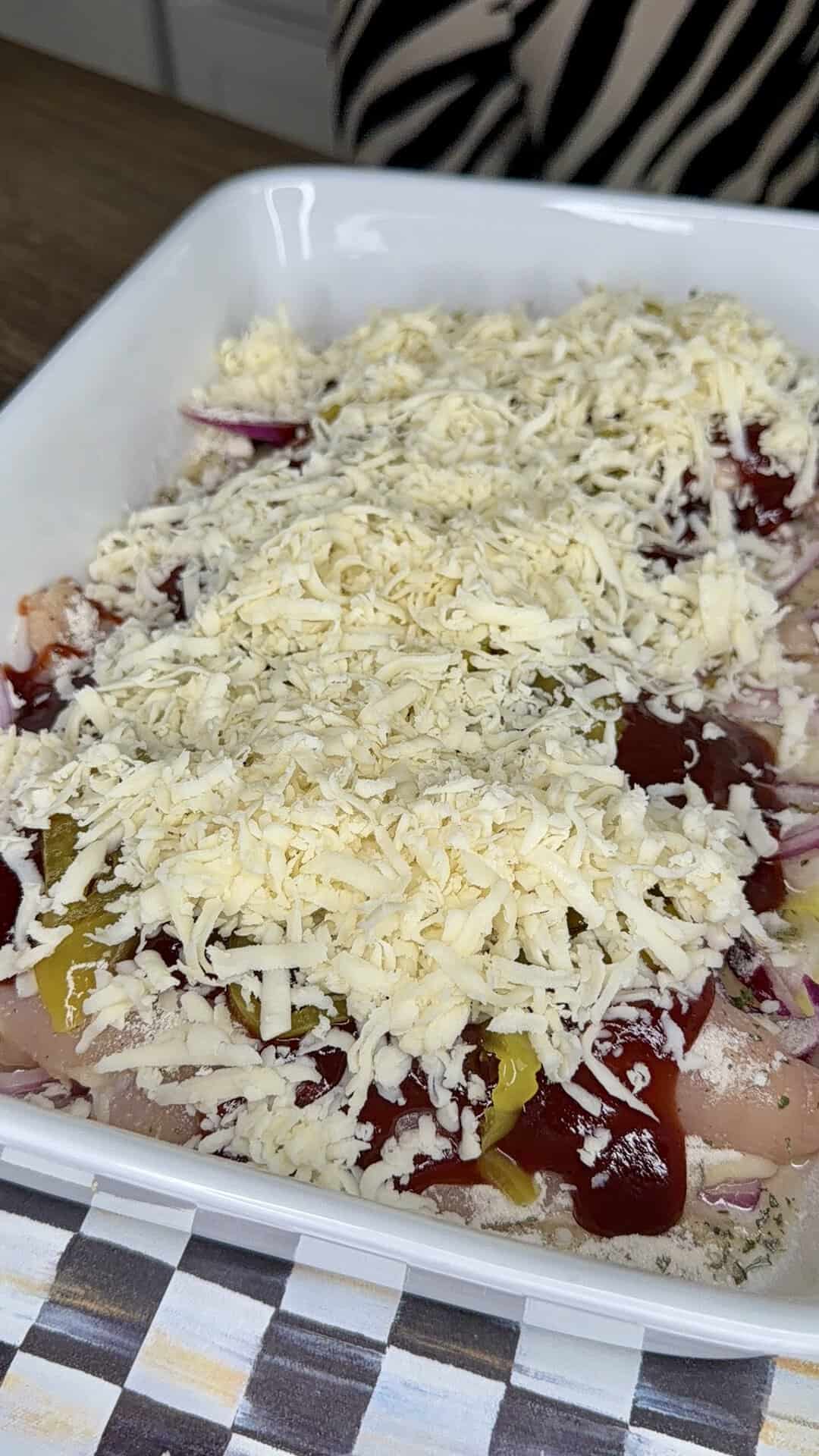 A white dish filled with raw chicken topped with shredded cheese, red onions, sliced peppers, and a red sauce, ready to be baked.