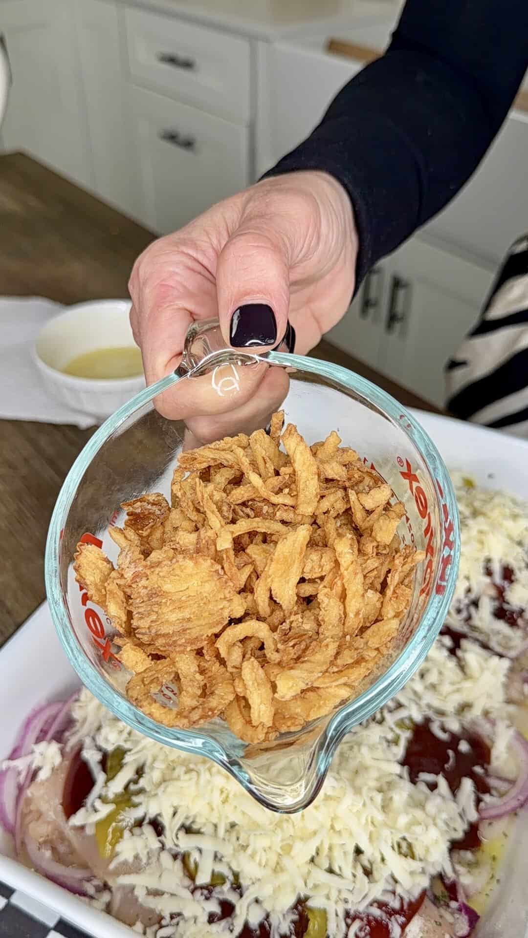 A hand holding a glass measuring cup filled with crispy fried onions over a baking dish with cheese and sliced vegetables, preparing to add the topping. Kitchen cabinets and a counter are visible in the background.