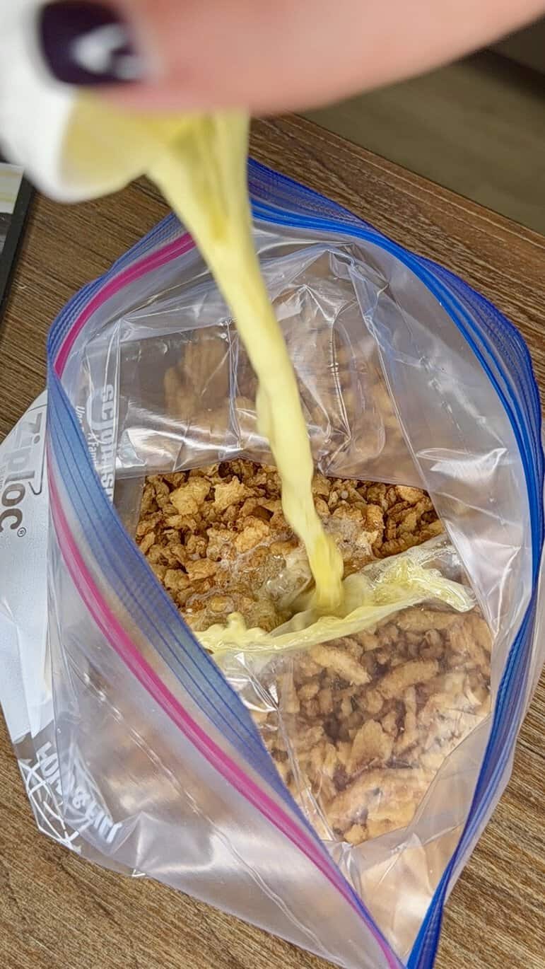 A hand squeezes a lemon, pouring juice onto granola inside a zip-top plastic bag on a wooden surface.