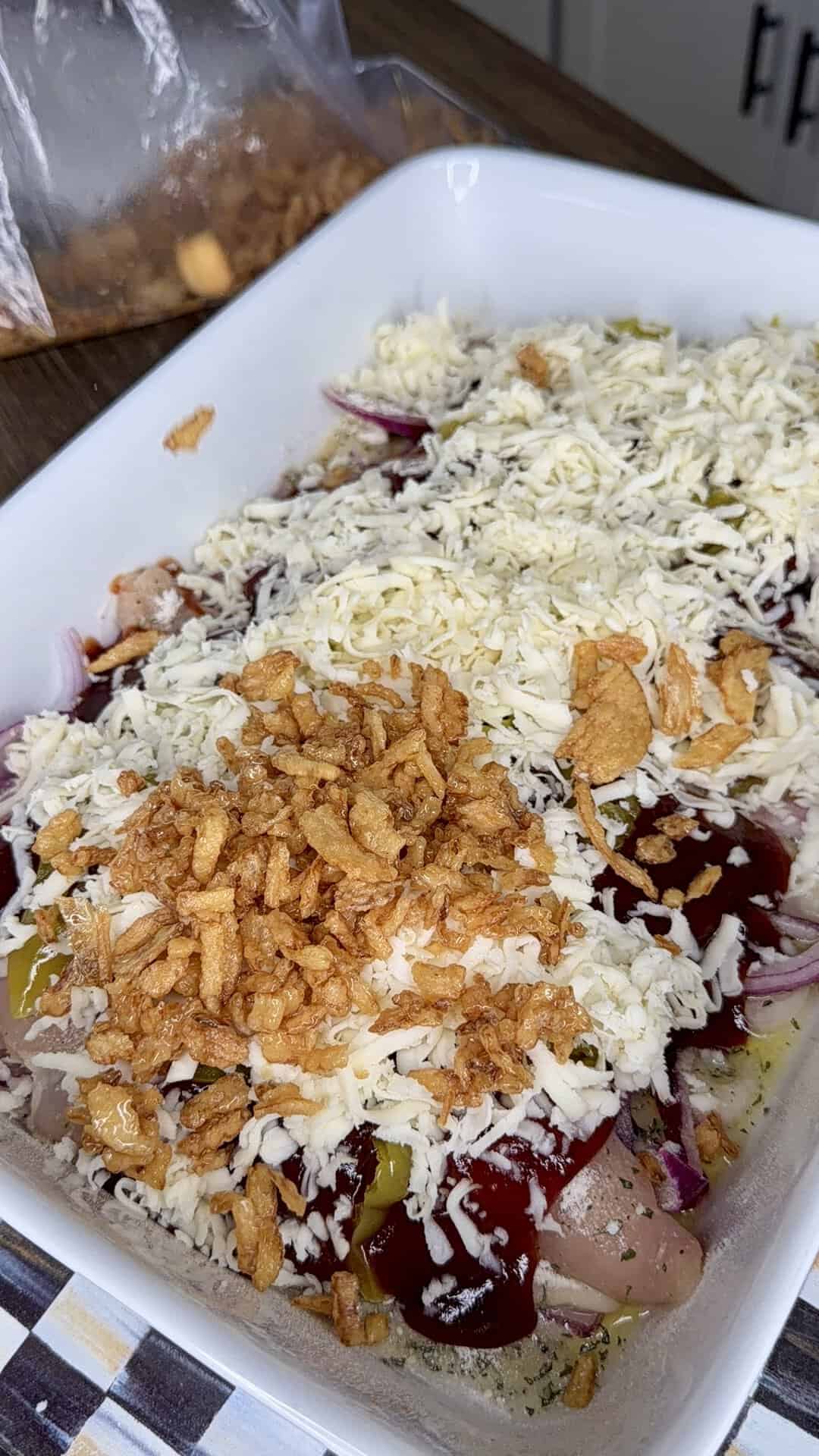 A white baking dish filled with raw chicken, shredded cheese, ketchup, sliced onions, and crispy fried onions, ready to be baked. A bag of fried onions is nearby on a wooden countertop.