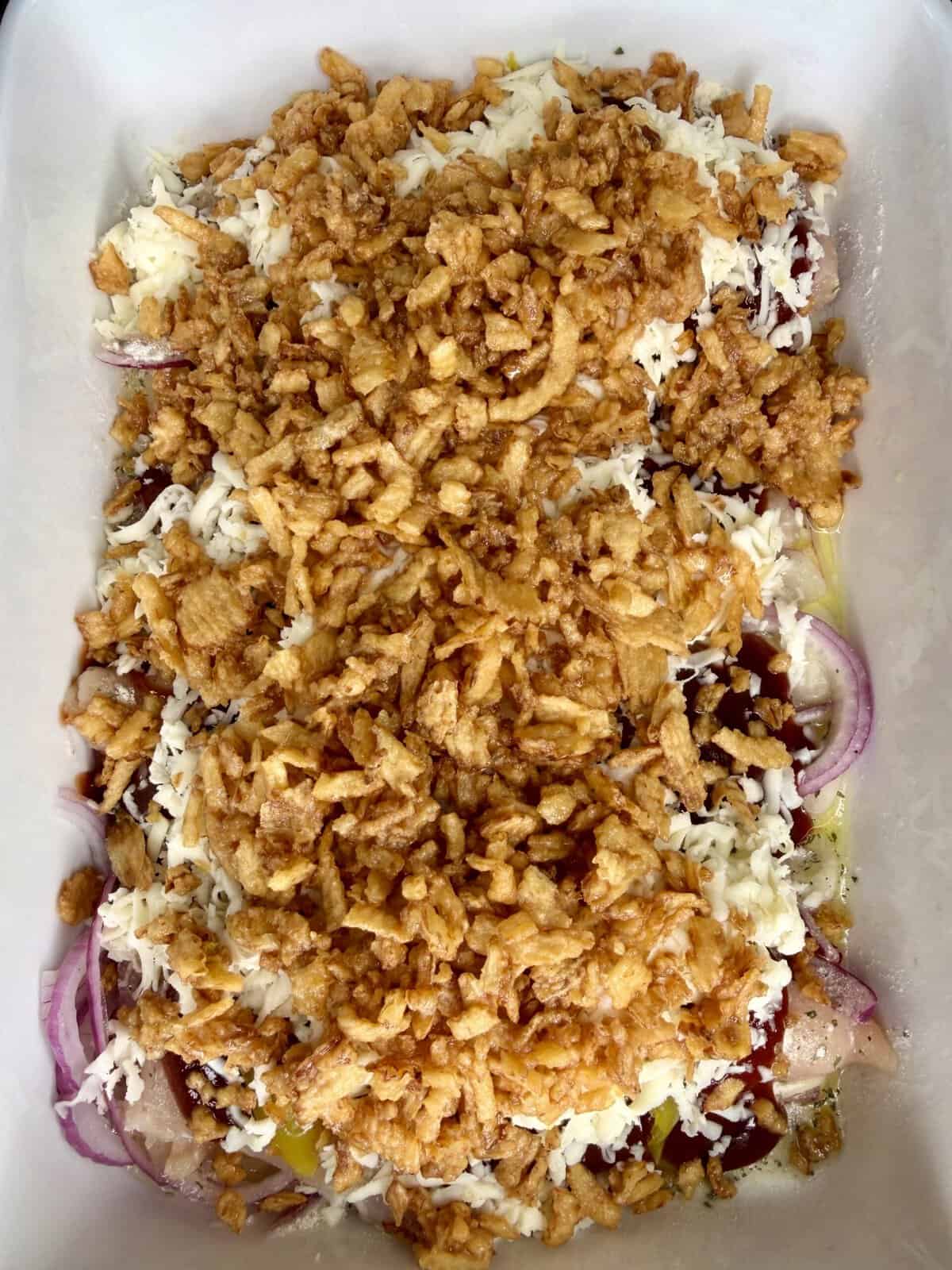 A casserole dish filled with layers of sliced red onions, shredded cheese, and topped with crispy fried onions, ready to be baked.