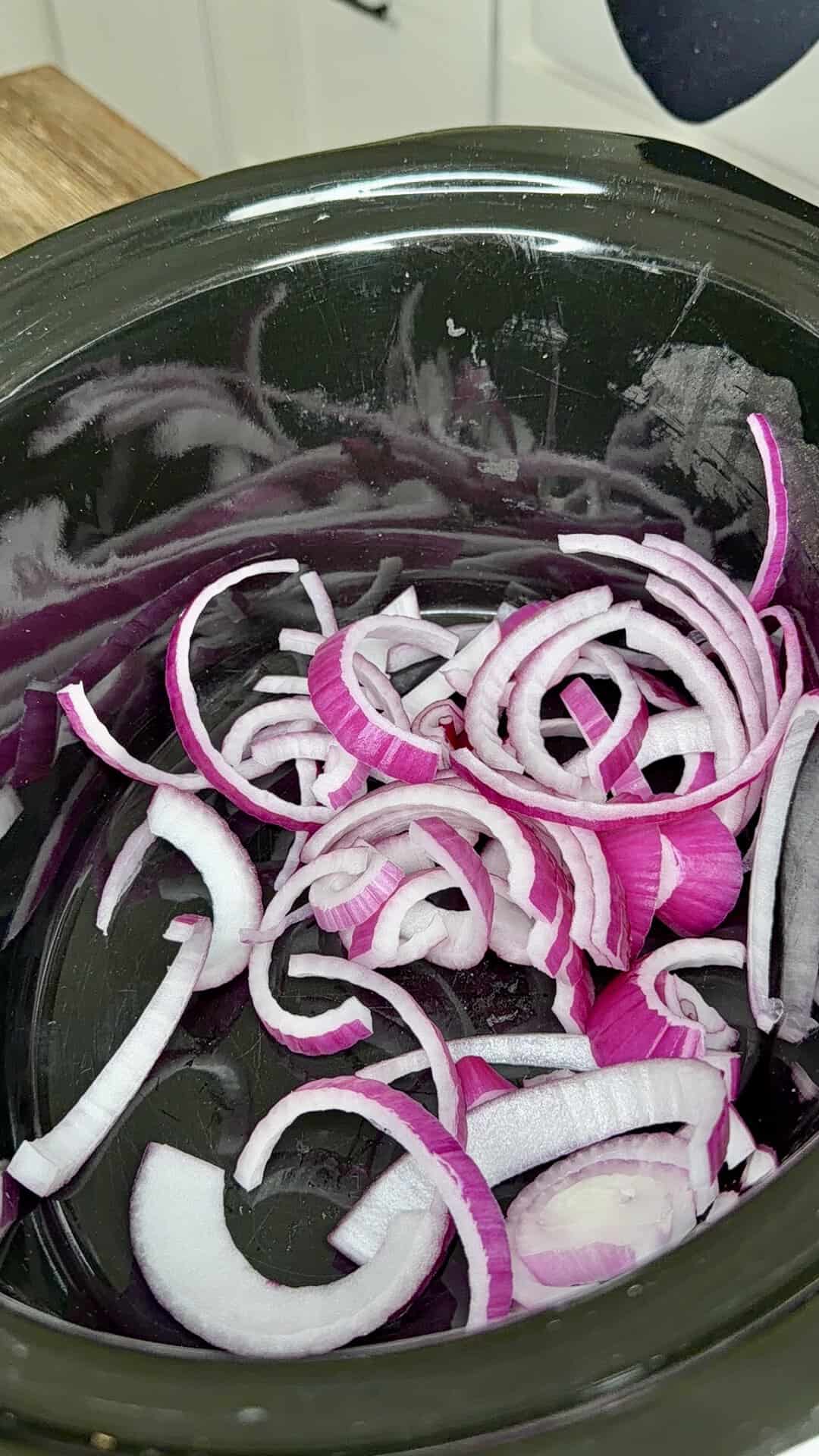 Sliced red onions in a black slow cooker, ready for cooking.