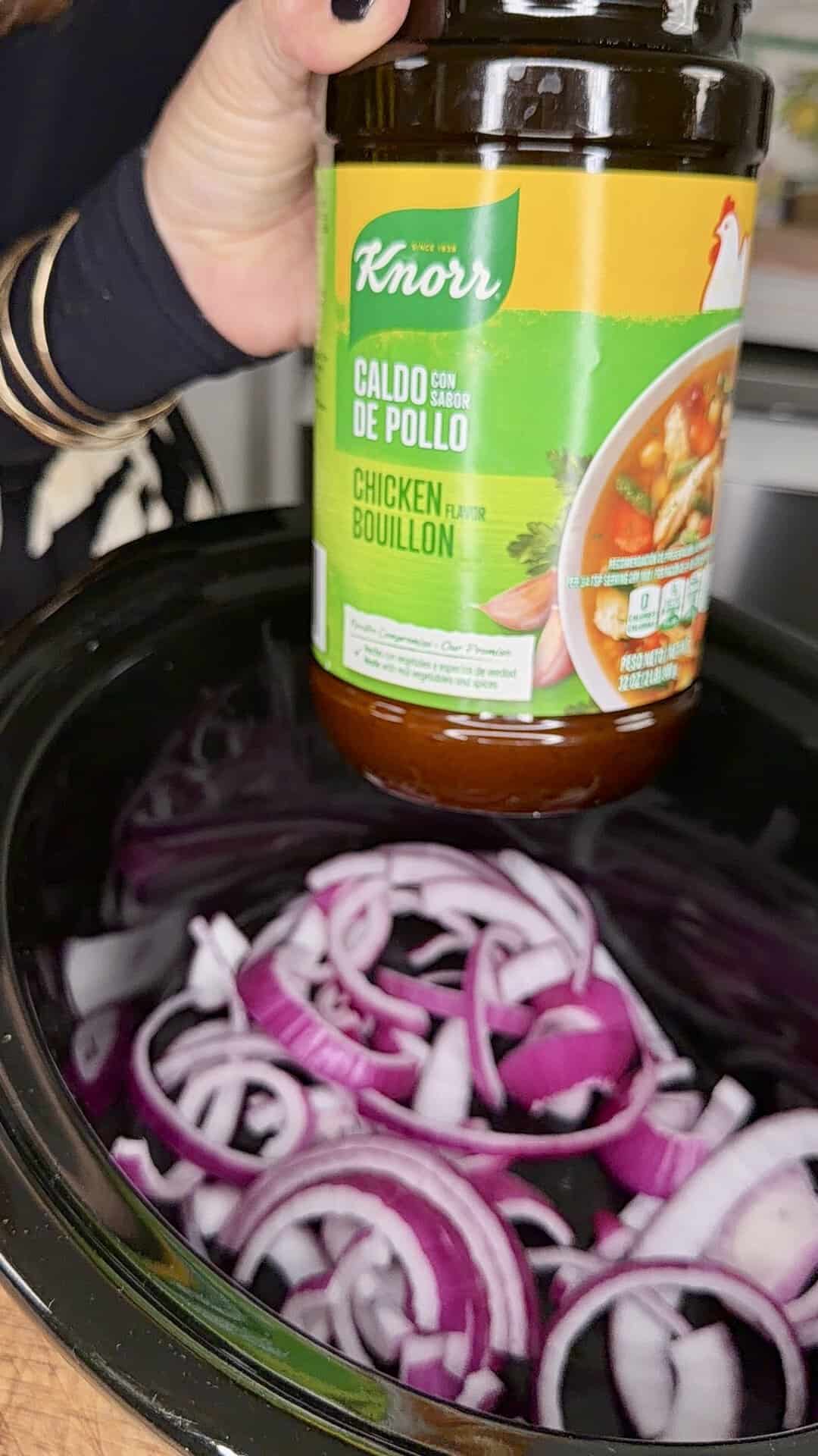 A hand holds a jar of Knorr chicken bouillon above a slow cooker filled with sliced red onions.