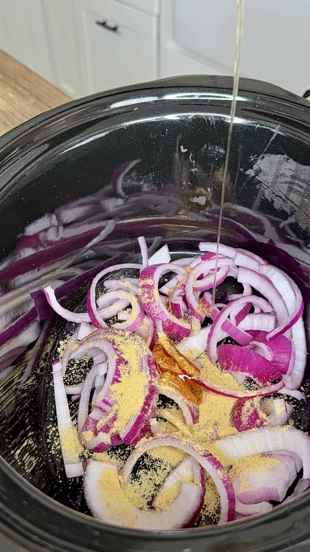 Sliced red onions with seasoning in a slow cooker, as oil is being poured over them. White kitchen cabinets are visible in the background.