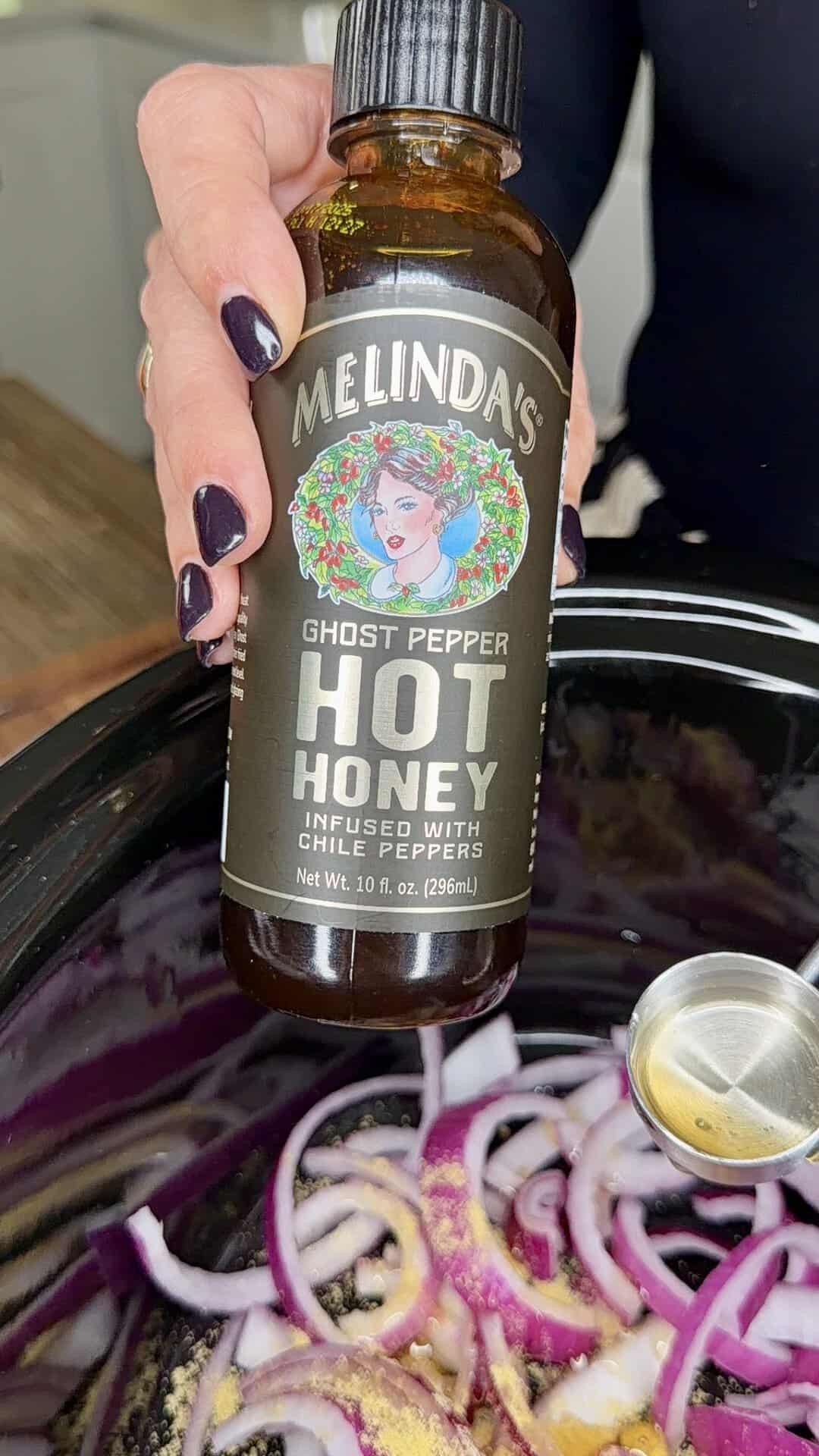 A hand holds a bottle of Melindas Ghost Pepper Hot Honey over a slow cooker filled with sliced red onions and a metal spoon.