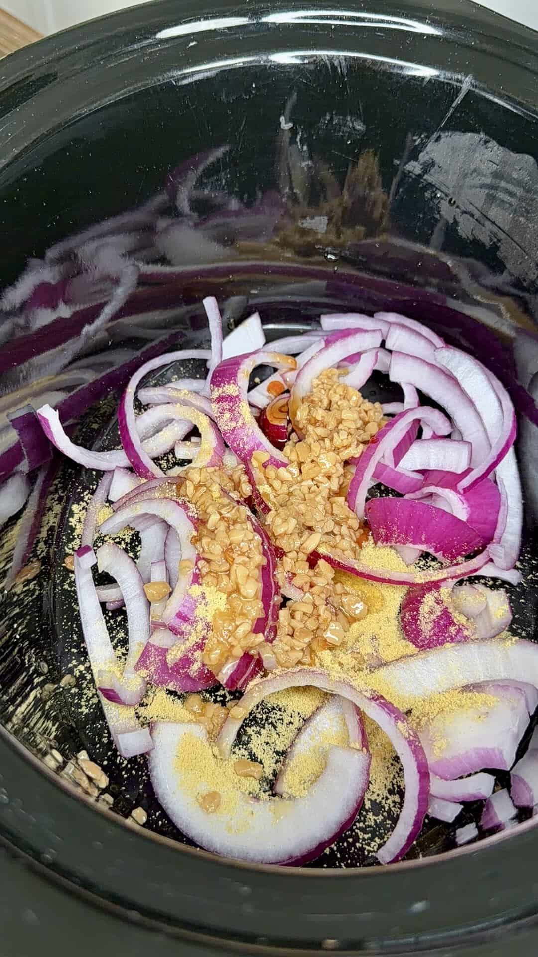 Sliced red onions, minced garlic, and yellow seasoning powder in a black slow cooker, ready for cooking.