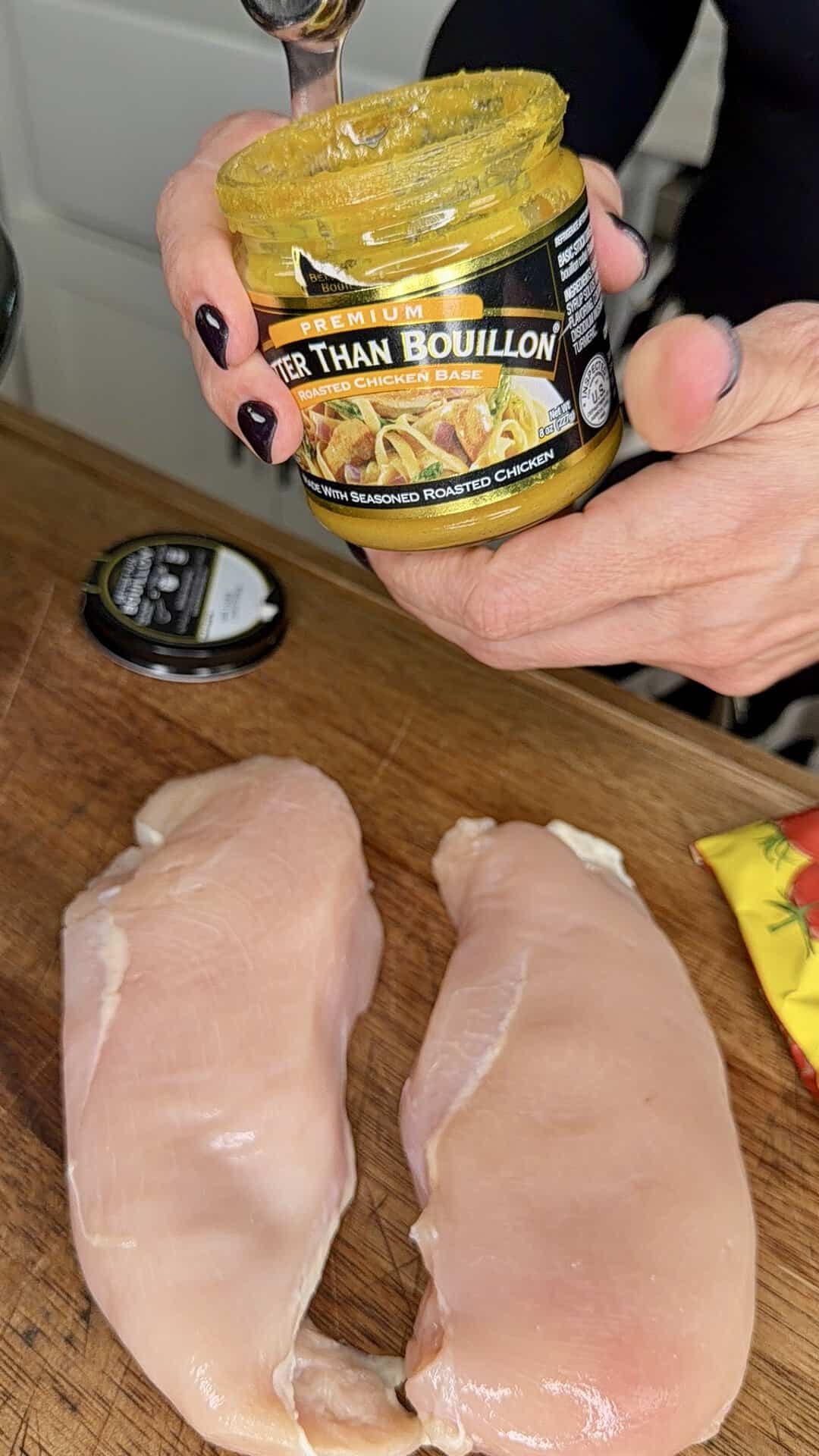 Two raw chicken breasts on a wooden cutting board with hands holding a jar of Better Than Bouillon roasted chicken base and a spoon.