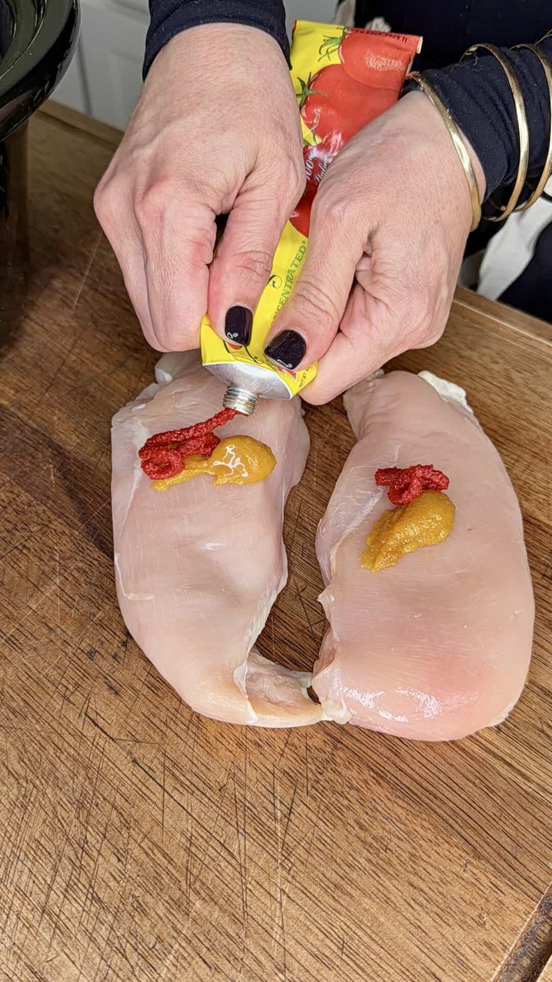 Two raw chicken breasts on a wooden board, each topped with tomato and mustard paste. A person with black nail polish squeezes a tube over the chicken.