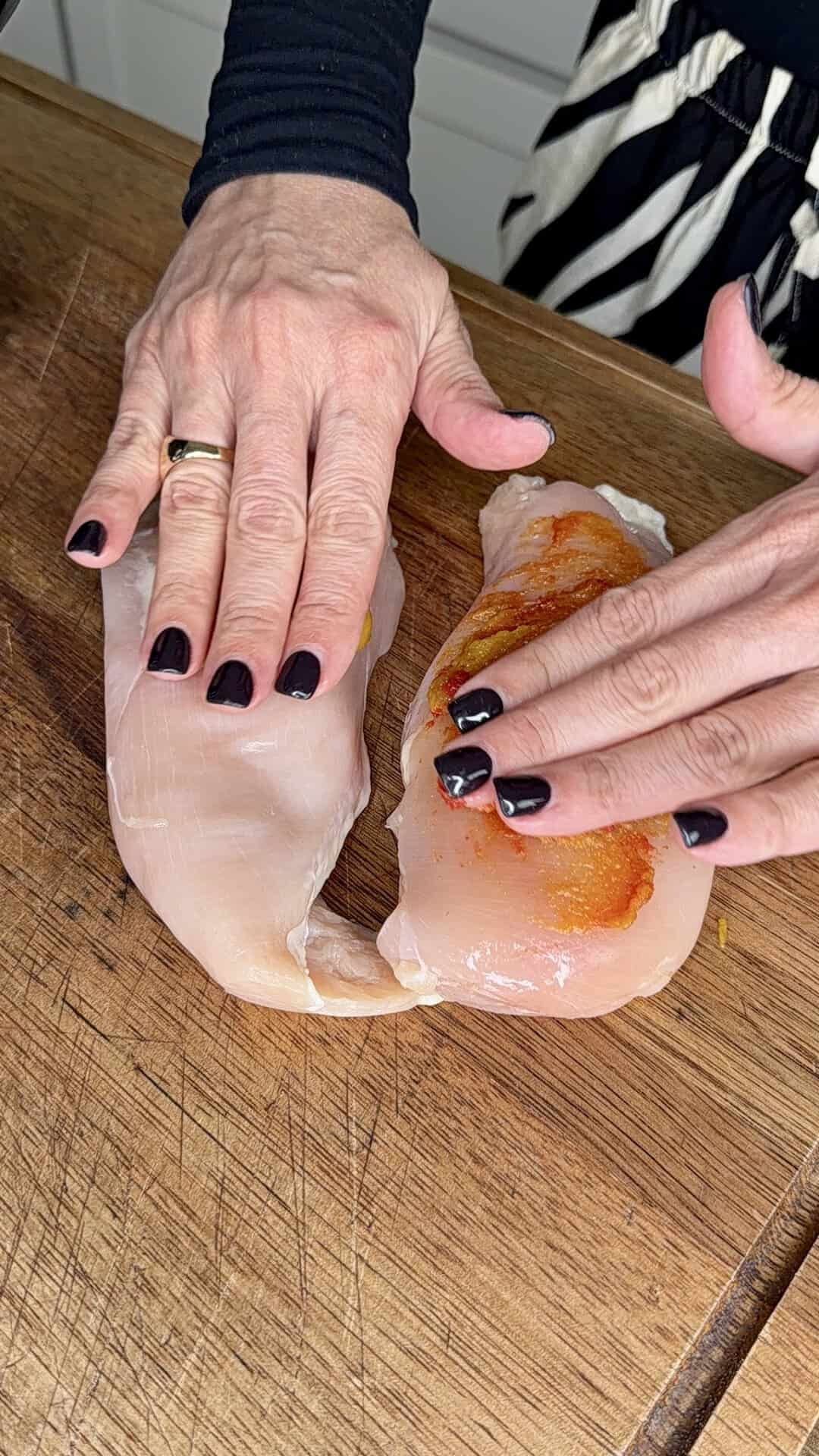 Two hands season raw chicken breasts on a wooden cutting board. The person has black nail polish and wears a gold ring on their left hand. Striped pants and a dark sleeve are partially visible in the background.